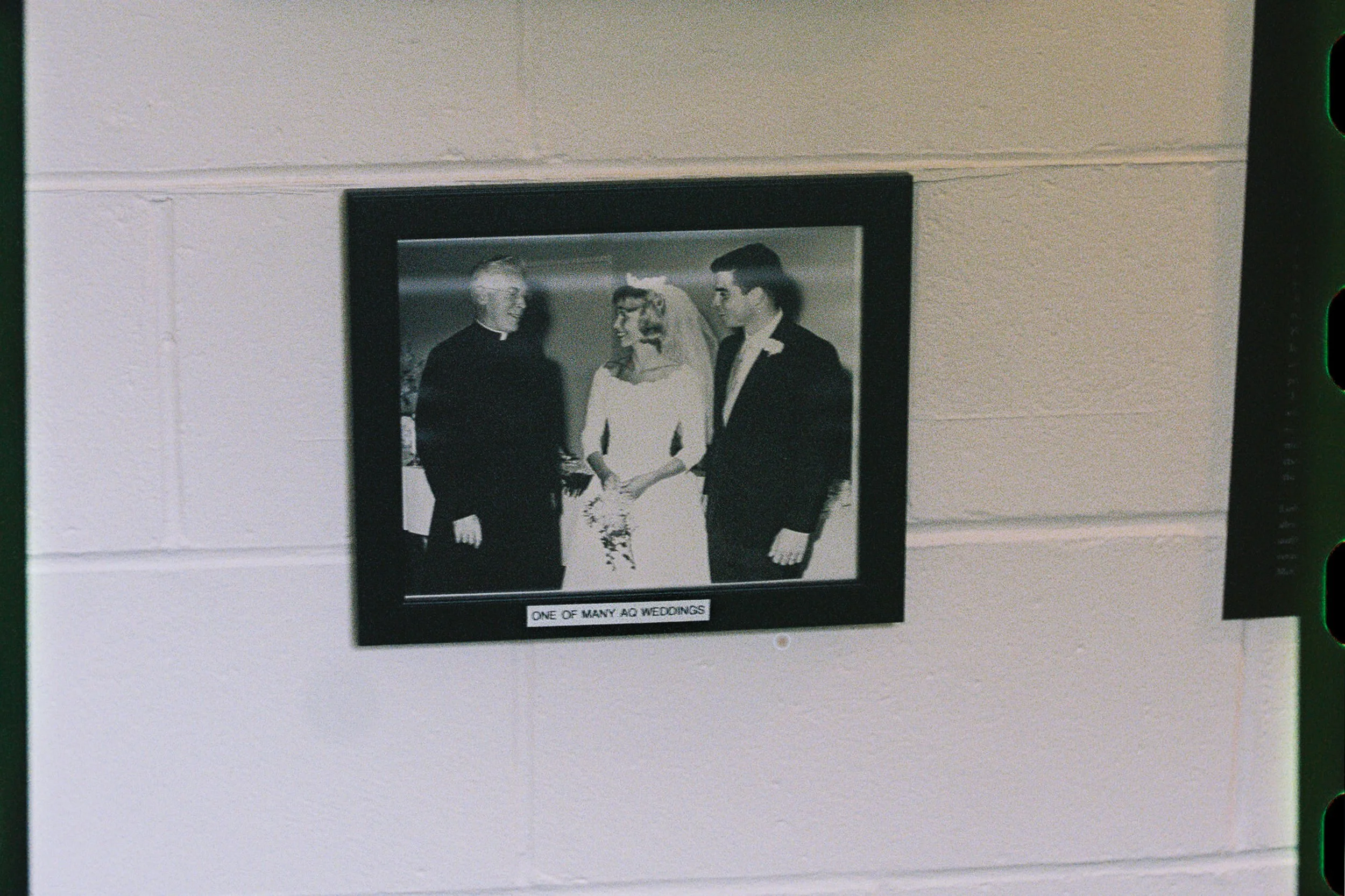 Black and white photo of a wedding with a woman in a wedding dress and veil, shaking hands with a man in a tuxedo. Another man in a tuxedo stands beside them, and a church or chapel can be seen in the background. Caption reads 'One of many AD wedding