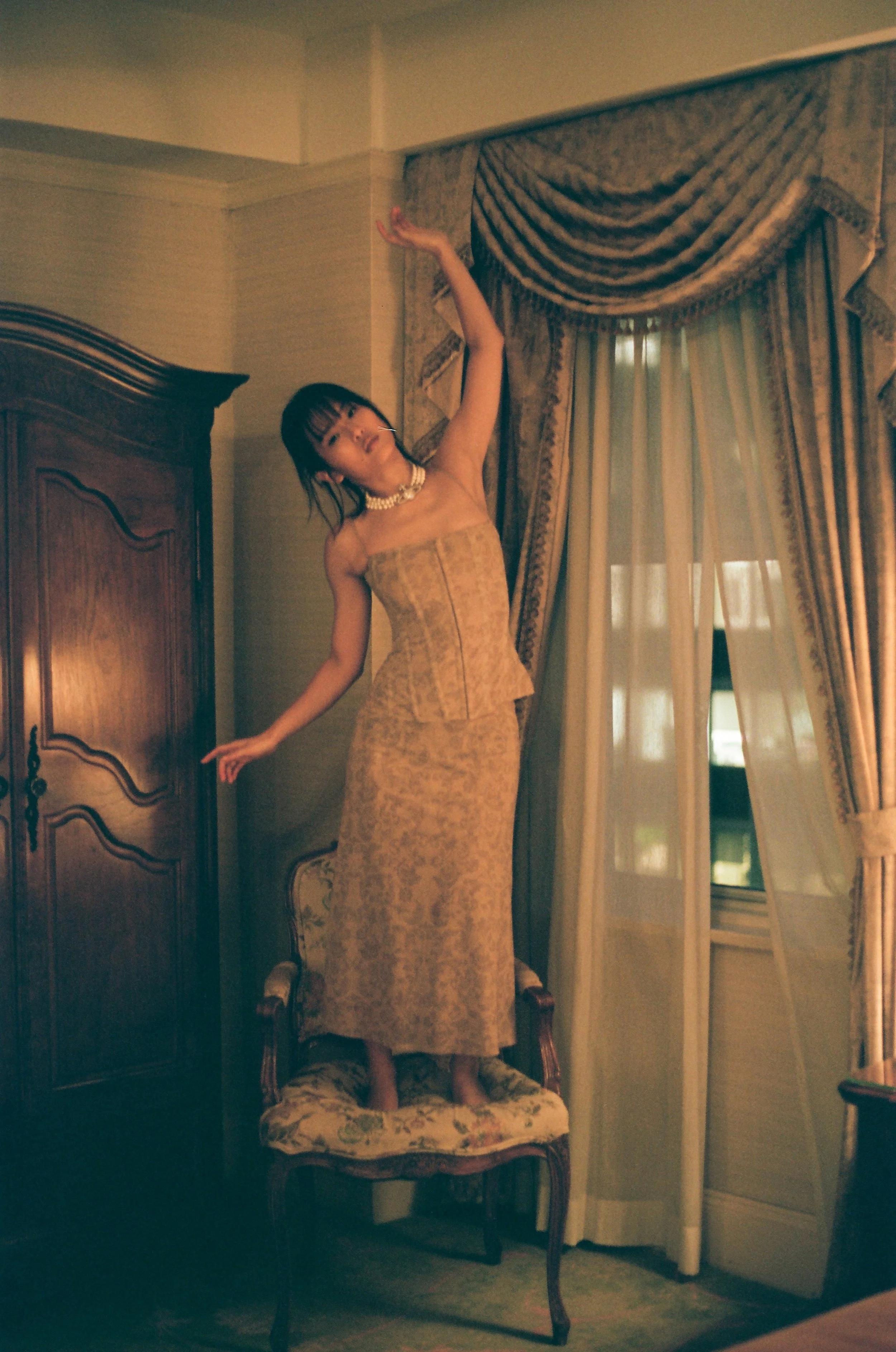 A woman standing on a vintage upholstered chair in a room with elegant draped curtains and antique furniture, striking a pose with arms raised.