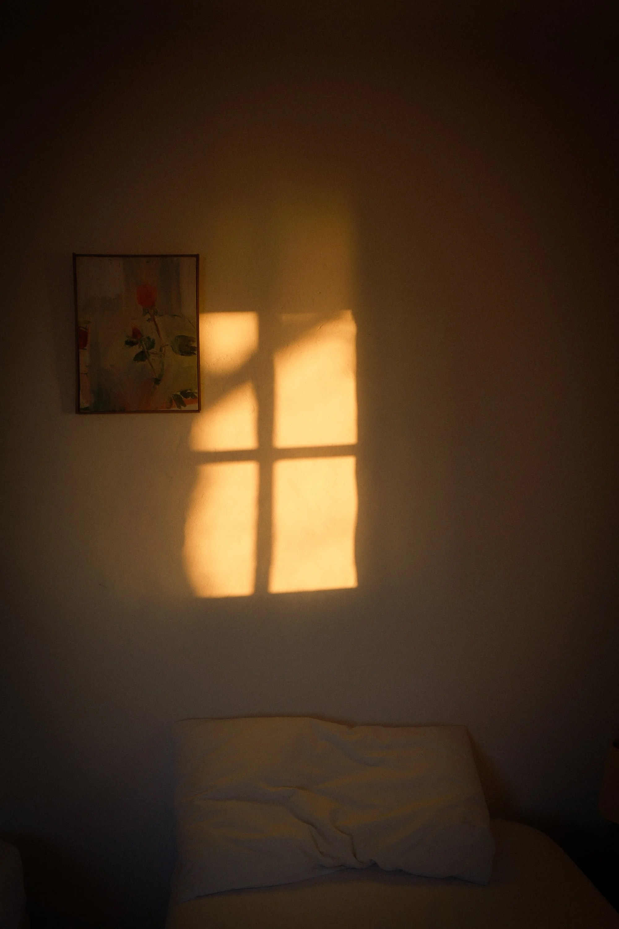 Sunlight coming through a window, casting a cross-shaped shadow on a plain wall, with a bed and pillow in the foreground.