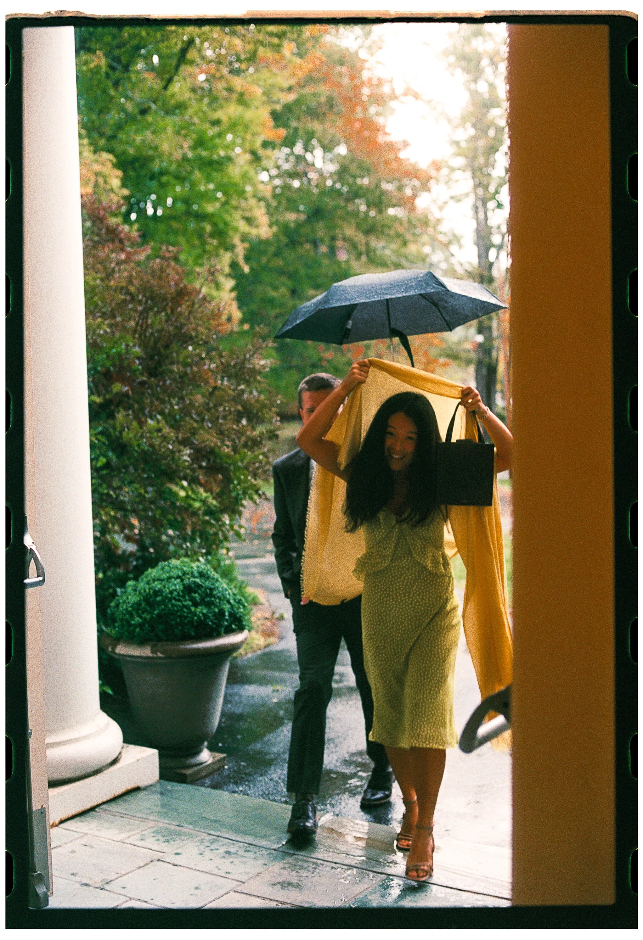 A woman with long dark hair wearing a yellow dress and heels is walking out in the rain holding a yellow raincoat over her head while a man holds an umbrella over her. They are outside near potted plants and trees, with the sun shining through the ba