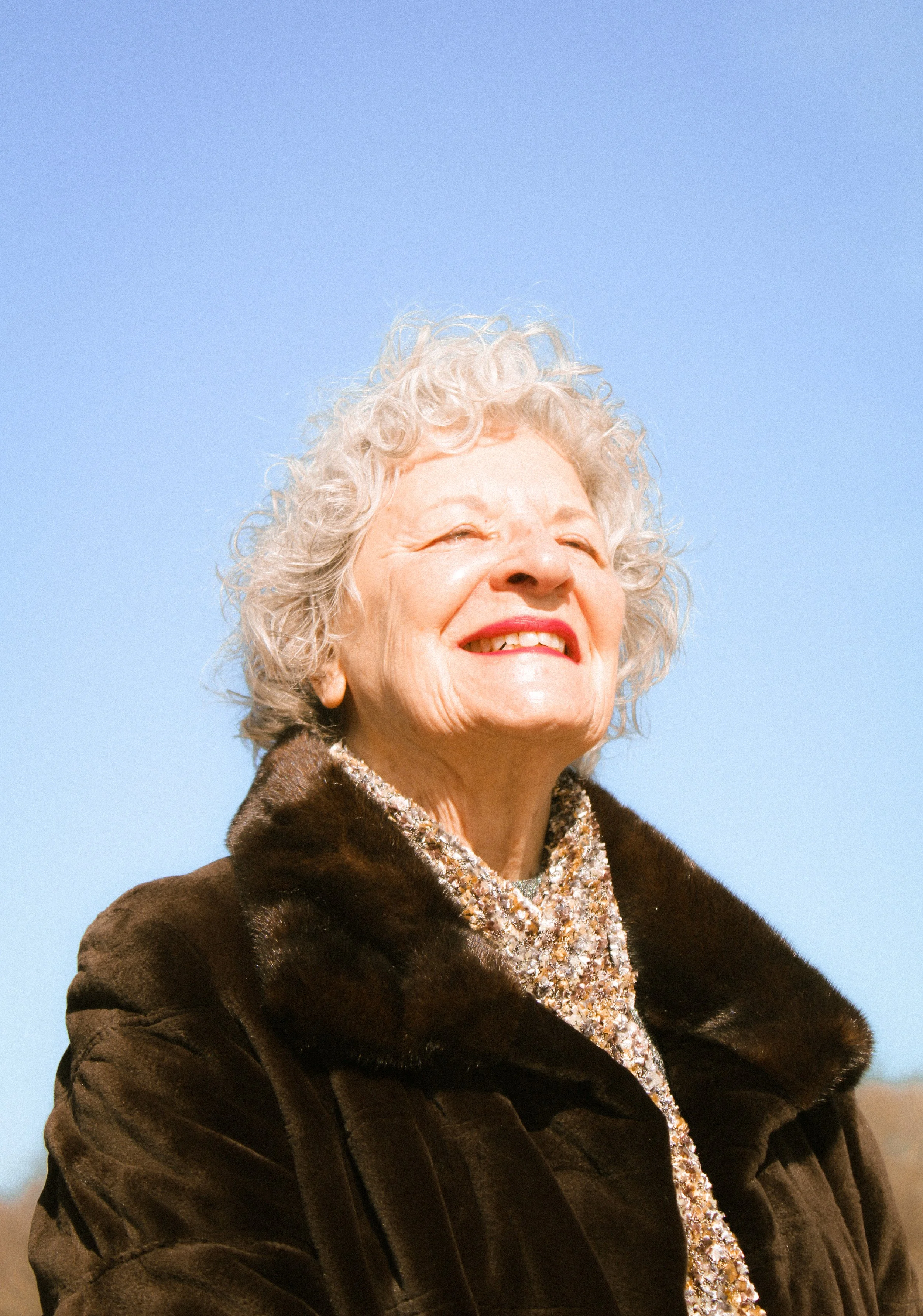 A smiling elderly woman with curly gray hair, wearing a dark brown fur coat and a sparkly silver dress, standing outdoors against a blue sky.