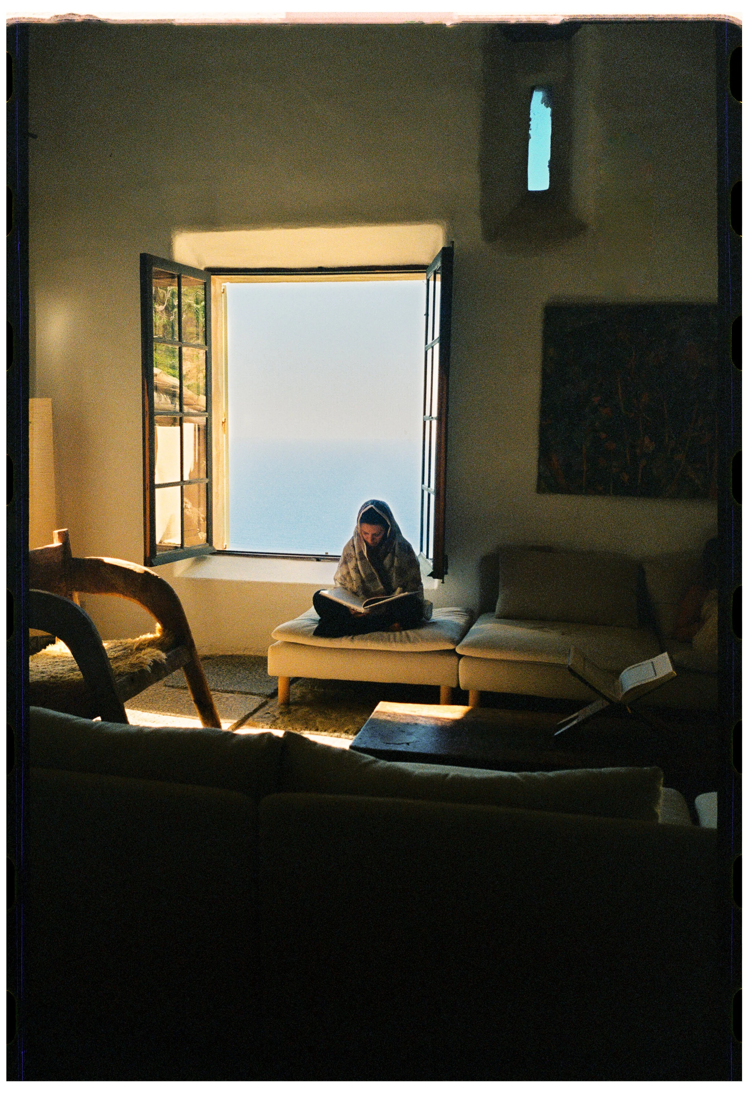 A woman sitting on a white ottoman, reading a book near an open window with blue sky visible outside, in a dimly lit room with a beige couch and an abstract painting on the wall.