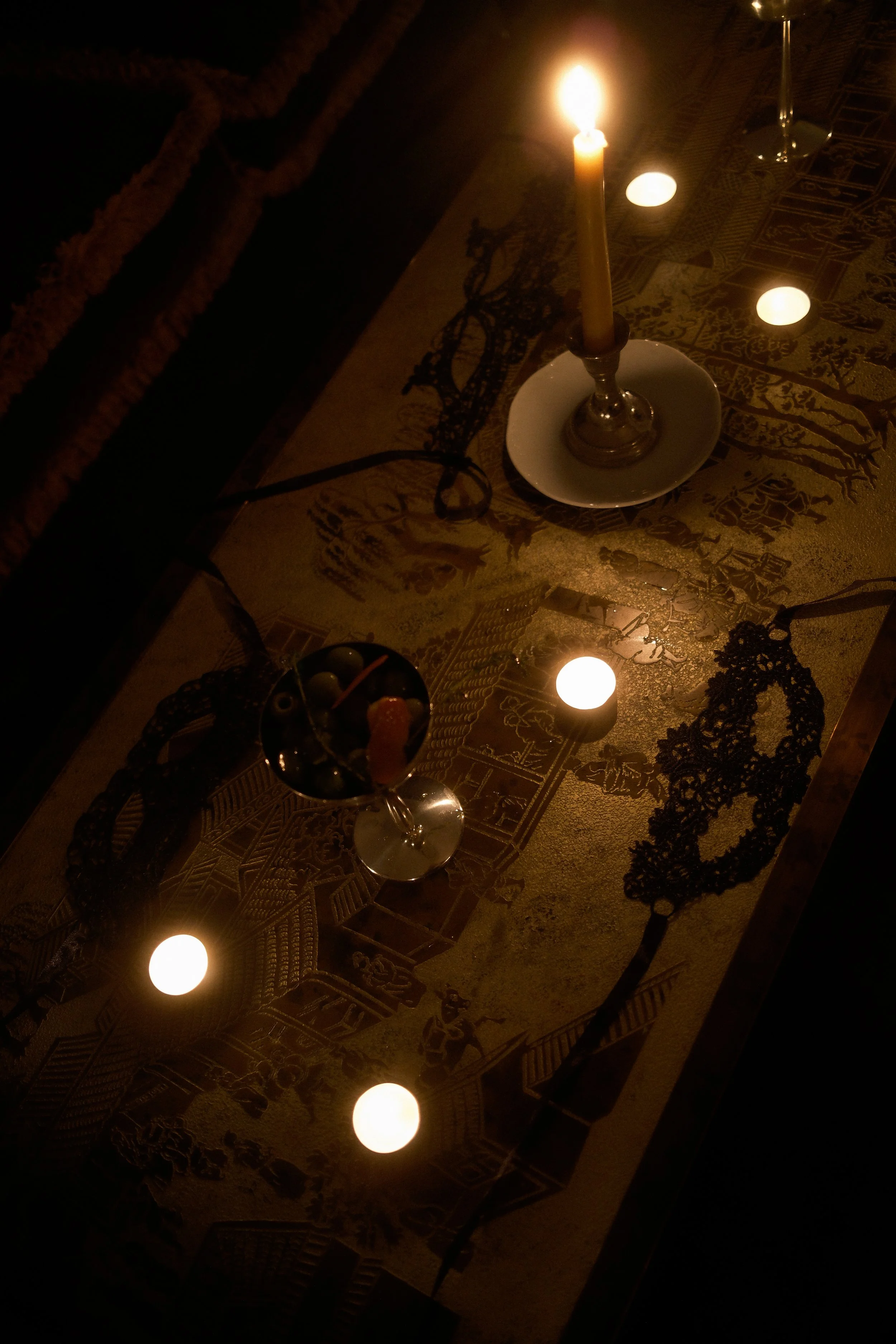 A table with a lit candle in a brass holder, surrounded by small candles and decorations, with a decorative cloth featuring Egyptian hieroglyphs and artifacts.