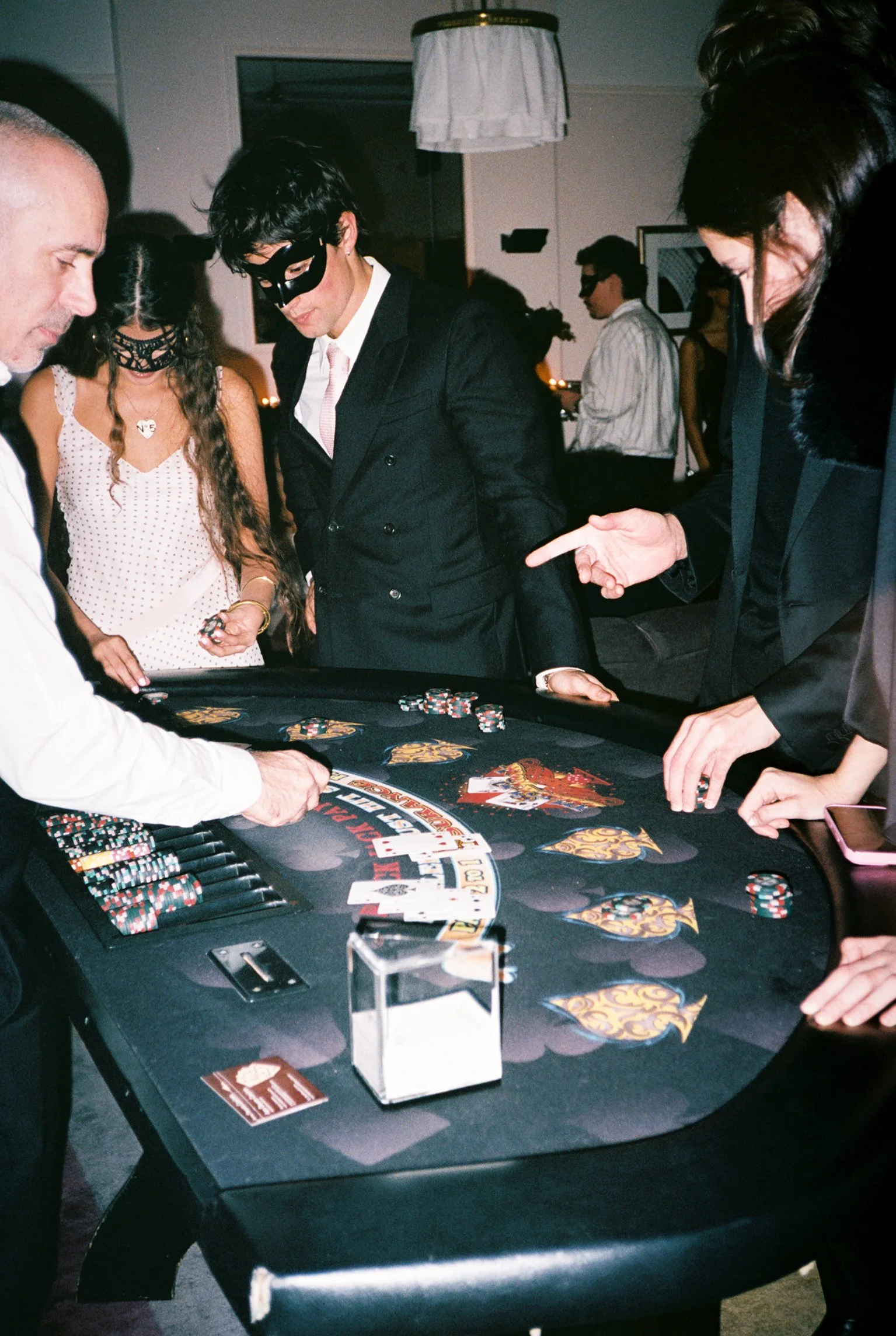 People playing poker at a casino table with chips, cards, and a dealer, wearing masks, in a dimly lit room.