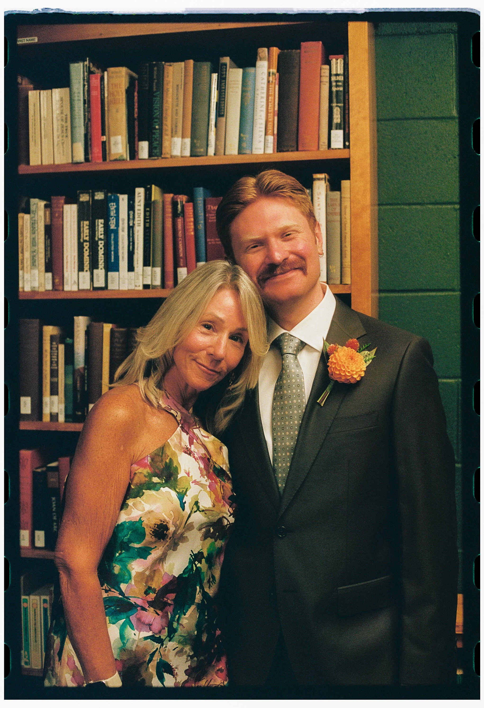 A man and woman smiling and posing together in front of a bookshelf filled with books. The woman has blonde hair and is wearing a floral sleeveless dress. The man has a reddish beard and mustache, and is wearing a dark suit with a light-colored tie a