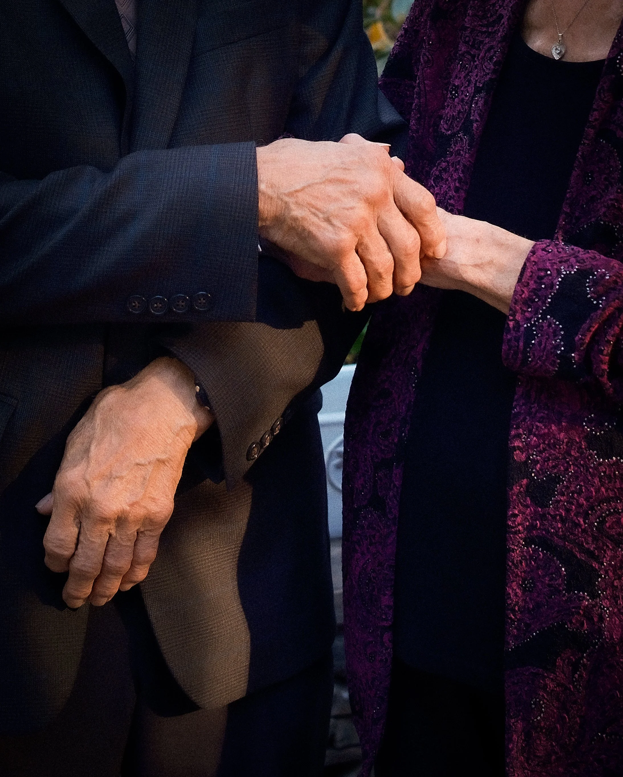 Close-up of two elderly individuals holding hands, one wearing a dark suit and the other in a purple patterned jacket. grandparents at an east coast wedding. Shot on 35mm film