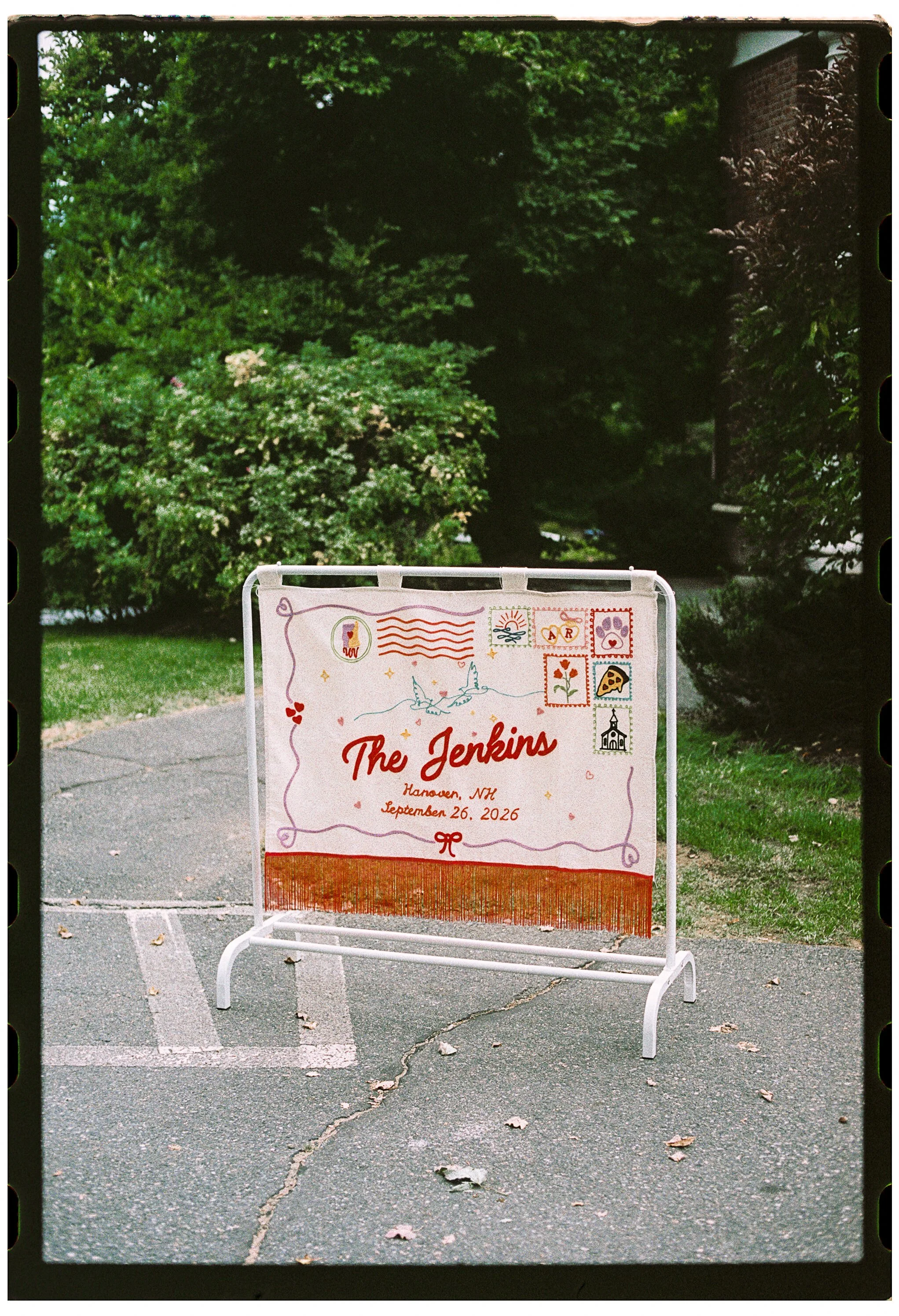 A decorative sign in a parking lot with the text 'The Jenkins, Hanover, NH, September 26, 2026,' surrounded by colorful illustrations of stamps, a unicorn, and a church, with green trees in the background.
