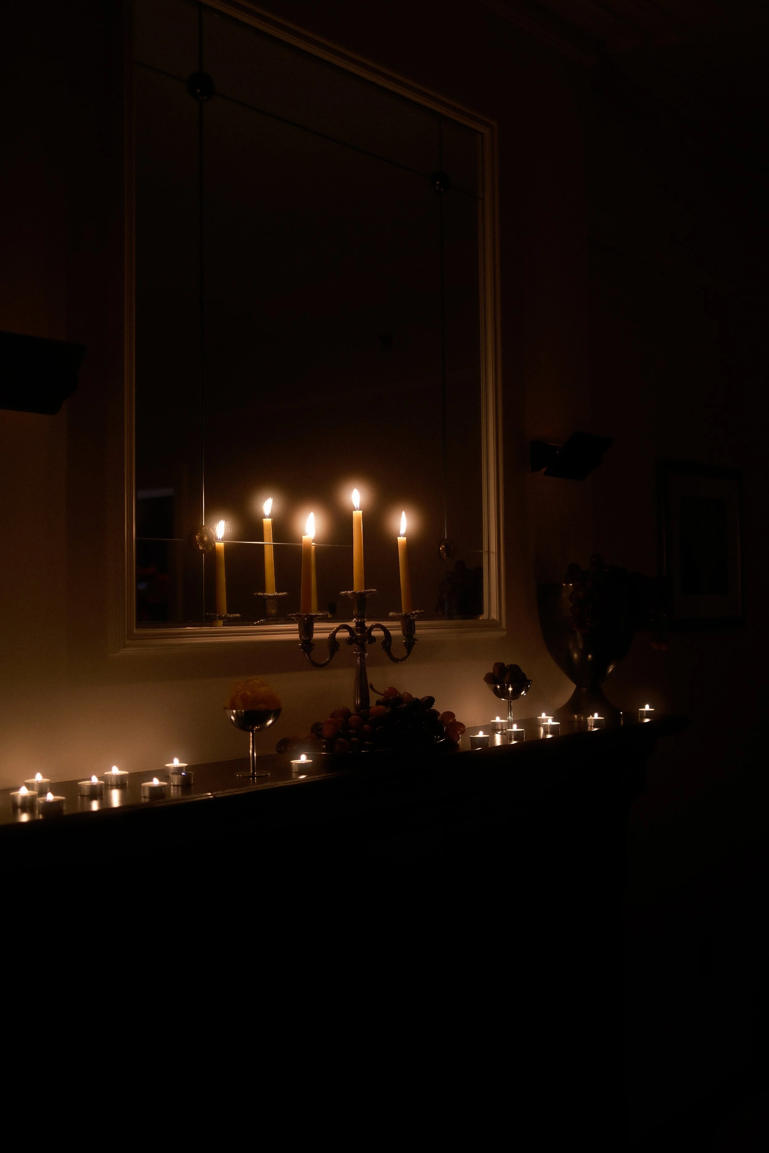 A mirror reflecting a room with a candelabrum holding five lit candles, a table with a large urn, small lit candles lining the edge of the table, and a bowl of fruit, with a framed picture on the wall and a dark indoor setting.