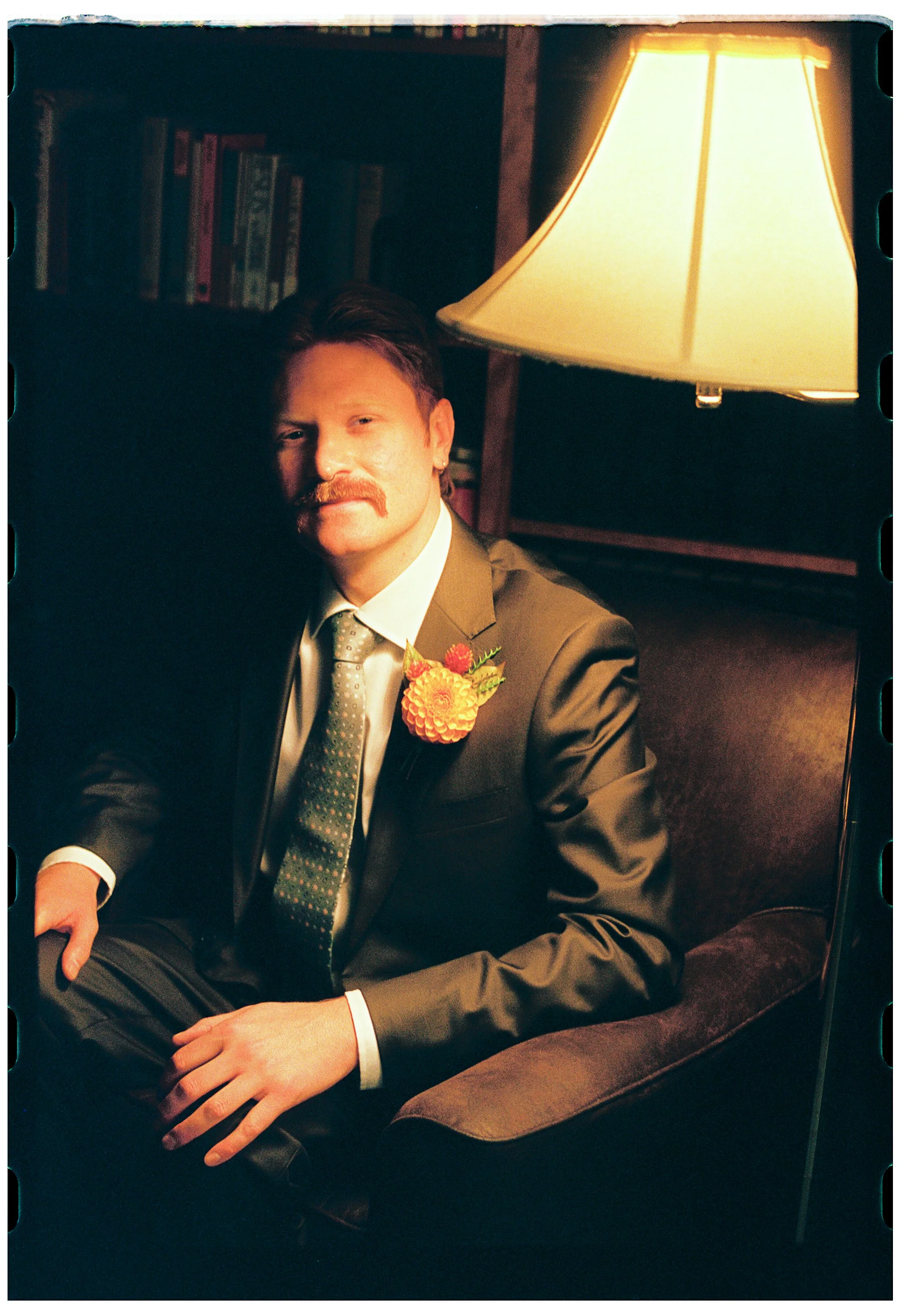 A man with a mustache and brown hair, dressed in a formal suit and tie, seated in a dimly lit room with a bookshelf and a large yellow lampshade behind him. He has a colorful boutonniere on his lapel and is looking at the camera.