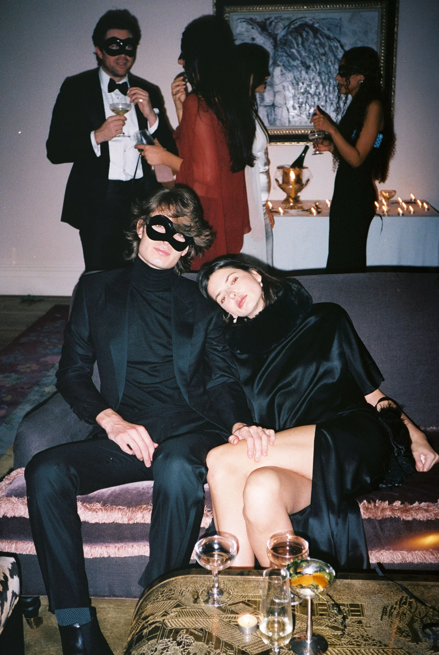 People at a party wearing masks, some holding drinks, with a woman lying on a man's lap in the foreground, in a decorated room with candles and artwork on the wall.