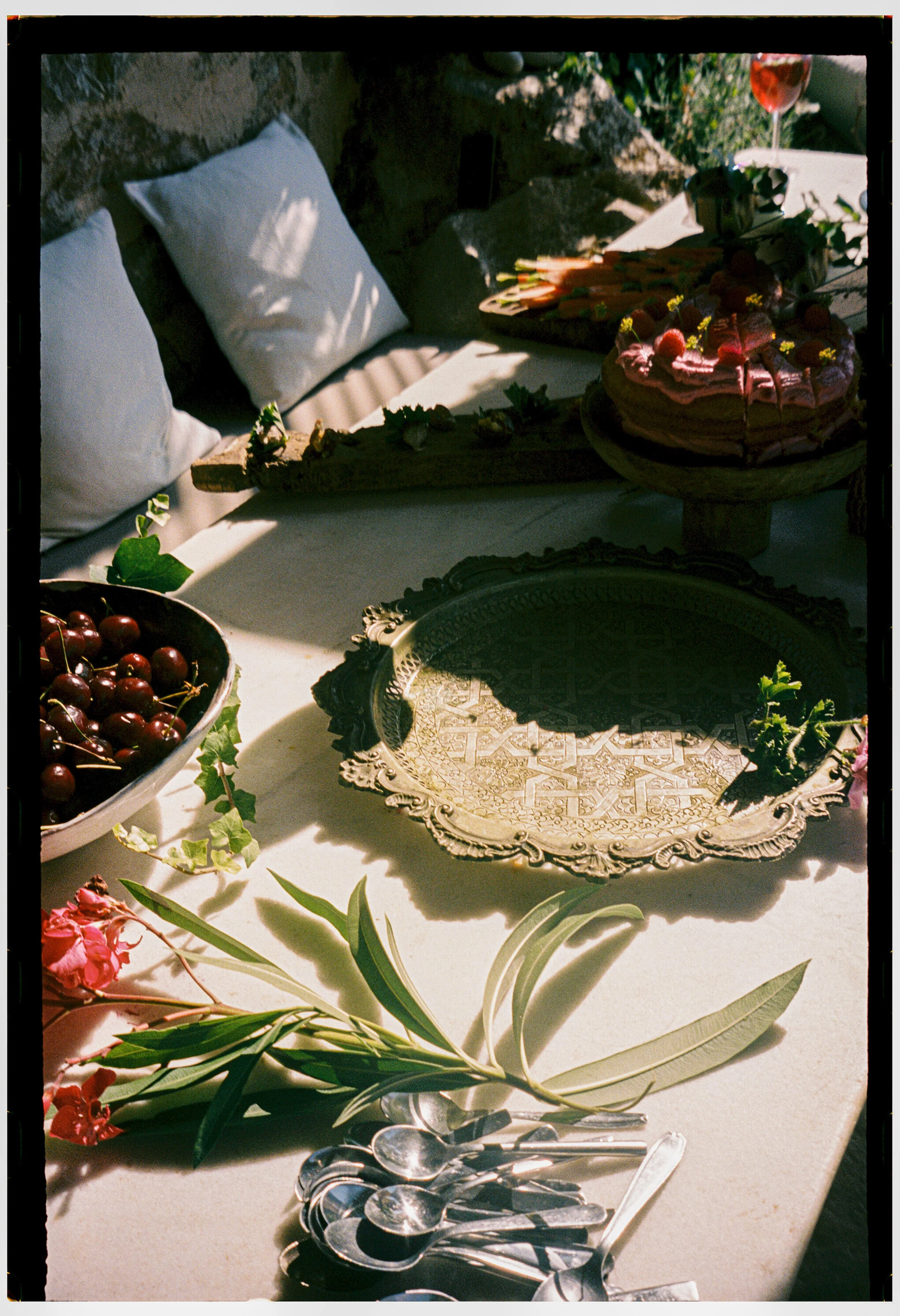 A table set with a cake topped with strawberries and pink frosting, a bowl of cherries, flower arrangements, and silverware, with white cushions and decorative plates in the background.