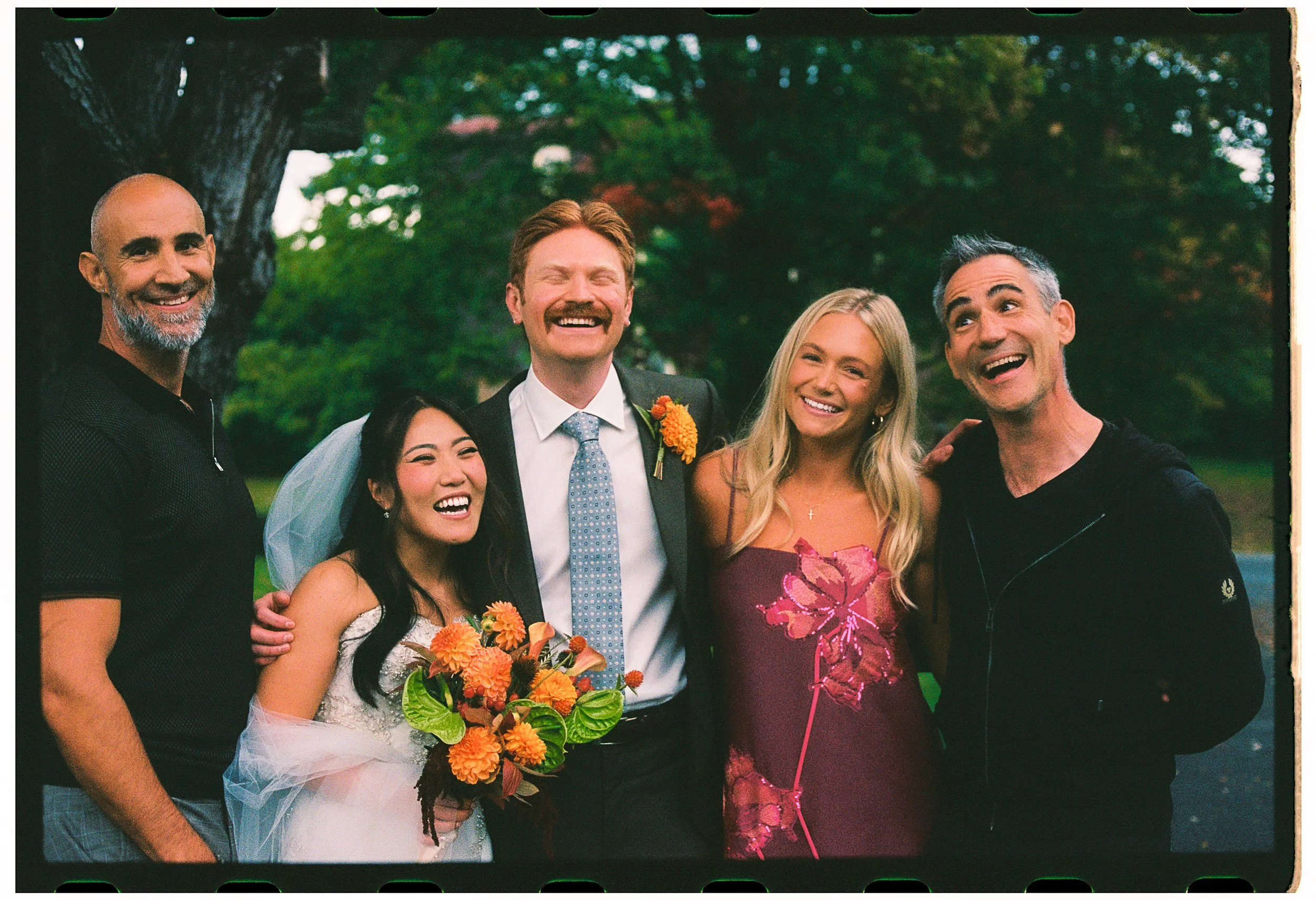 Group of six people smiling outdoors, with a green leafy background. Four women and two men, some with gray or dark hair, dressed in casual and semi-formal attire. One woman holds a bouquet of orange flowers.