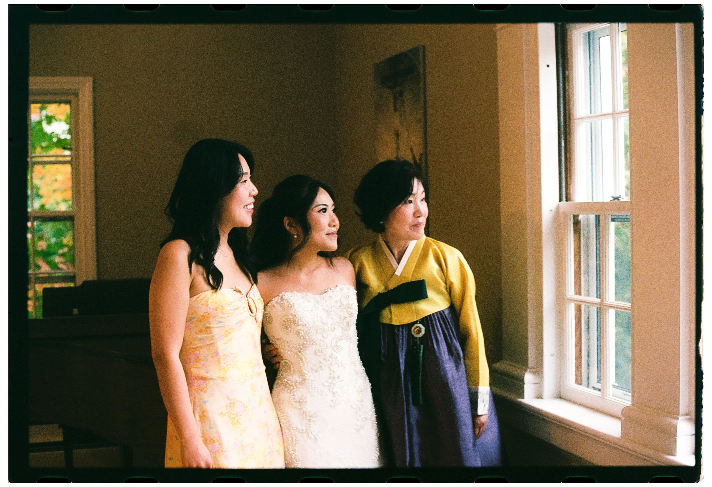 Three women smiling and standing by a window, wearing dresses and a hanbok, in a warmly lit room.