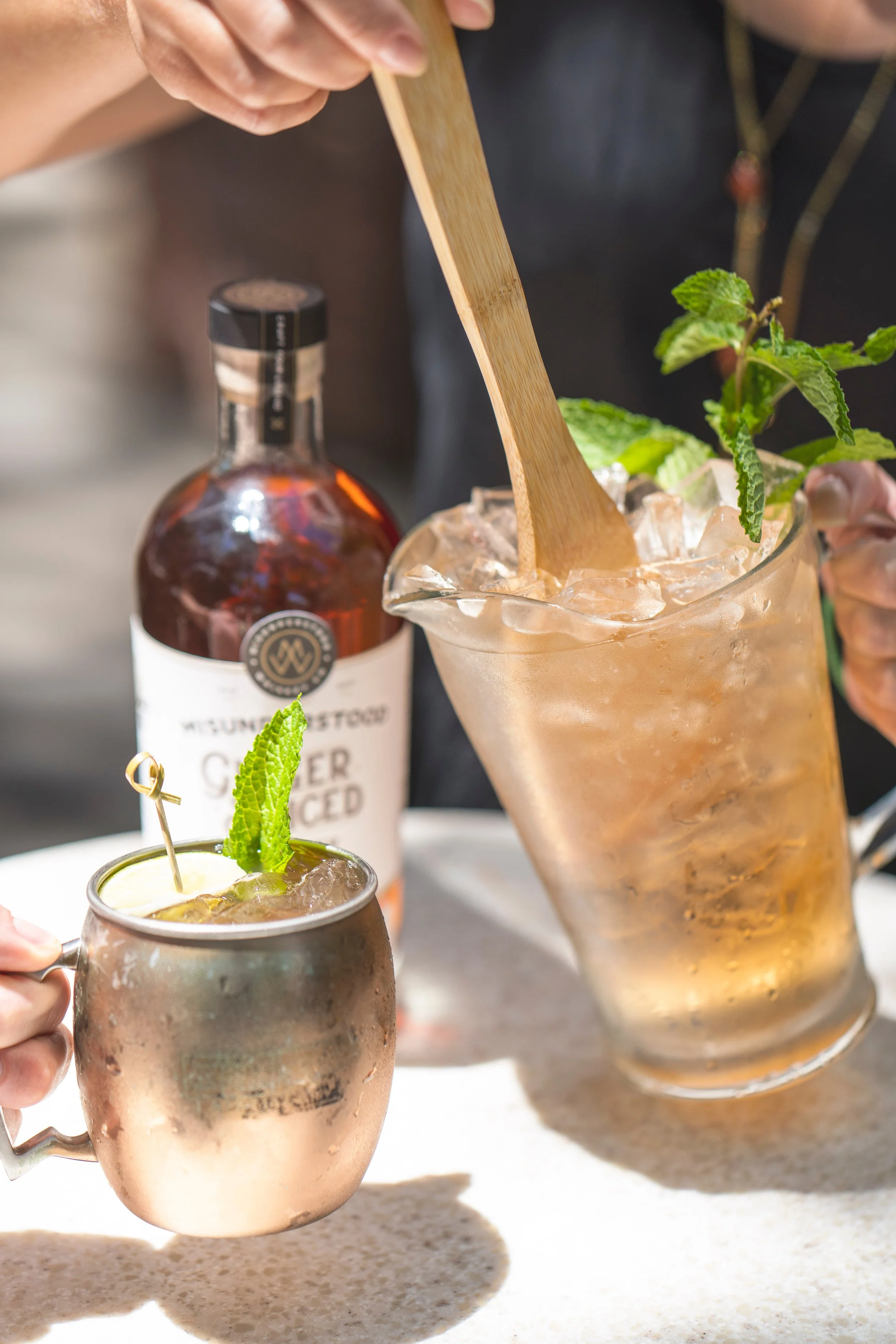 A person holding a copper mug filled with a cocktail garnished with a mint sprig, with ice inside and a lime wedge. In the background, a larger glass container with a similar cocktail and a sprig of mint, and a bottle of liquor. The scene appears to be at a bar or social gathering.