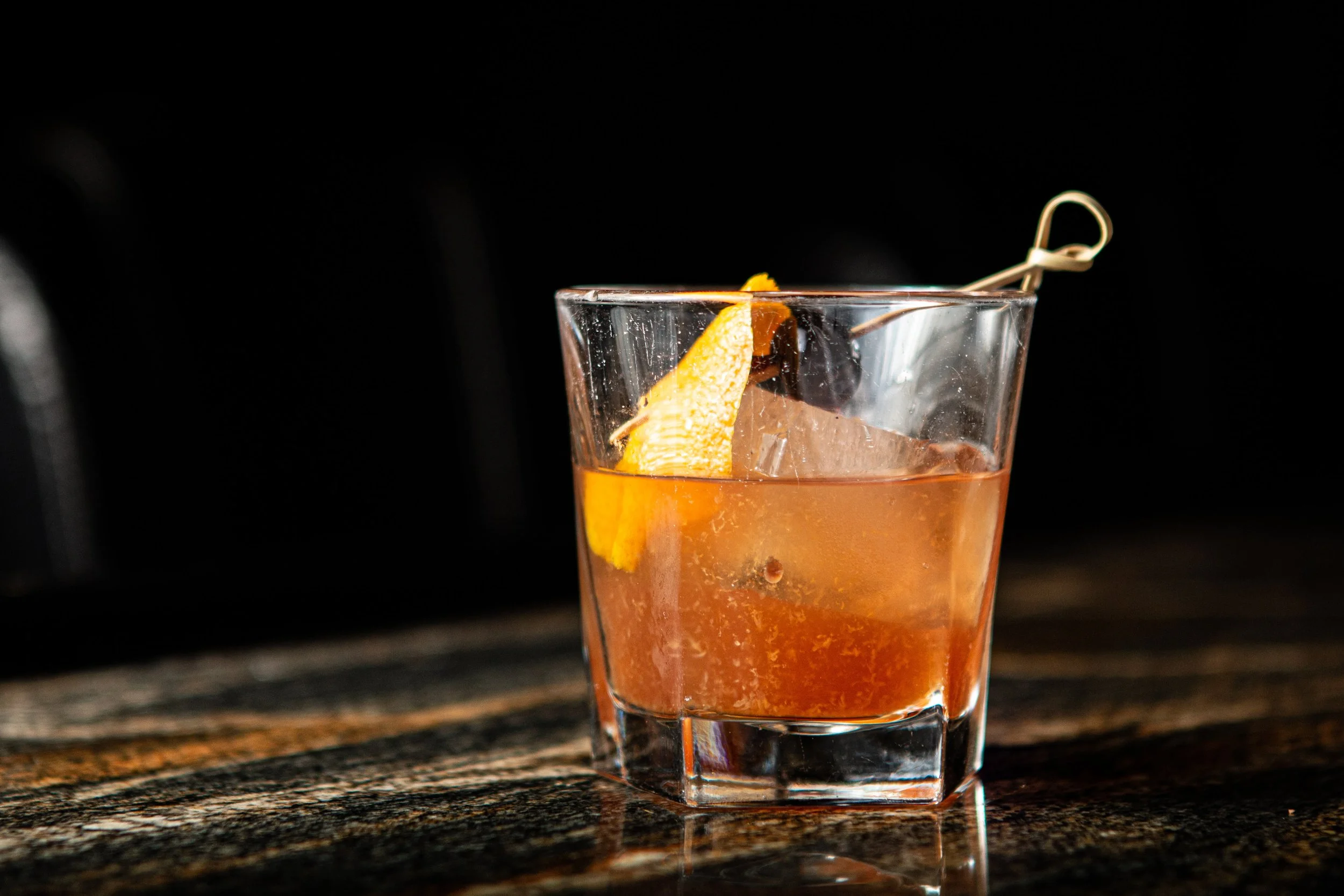 A glass of pinkish-orange drink garnished with an orange peel and a dark cherry, placed on a wooden surface against a black background.