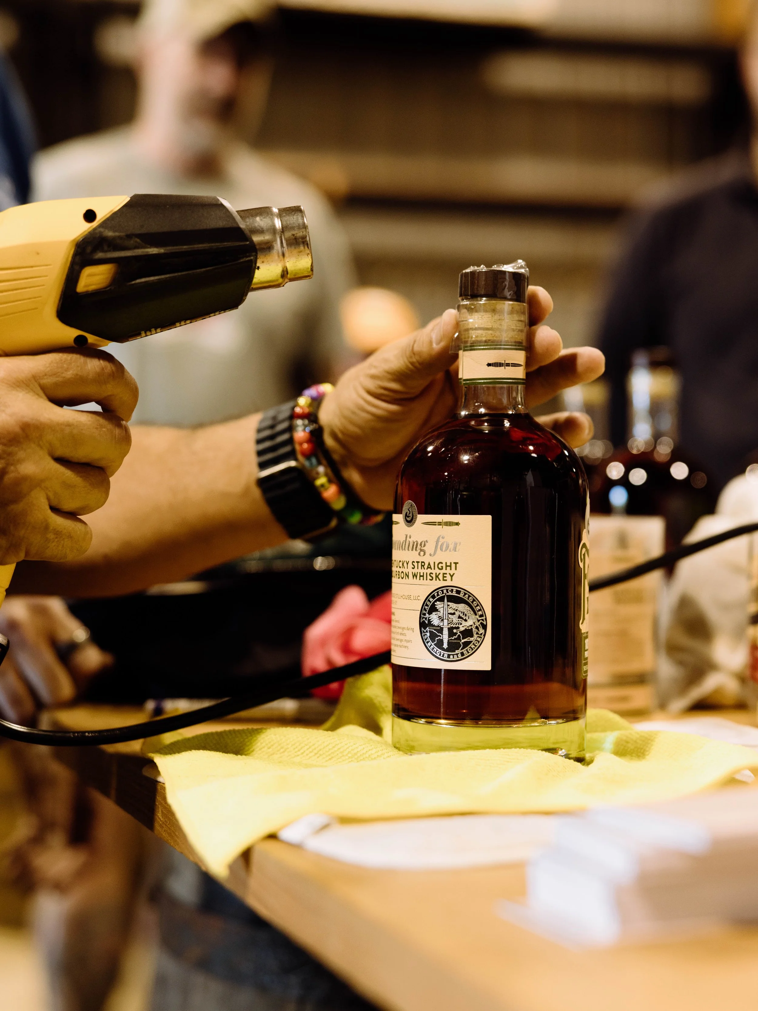 A person with a beaded bracelet is using a handheld heat tool on a bottle of whiskey, which is on a yellow cloth on a wooden surface. The background is blurry with other people around.