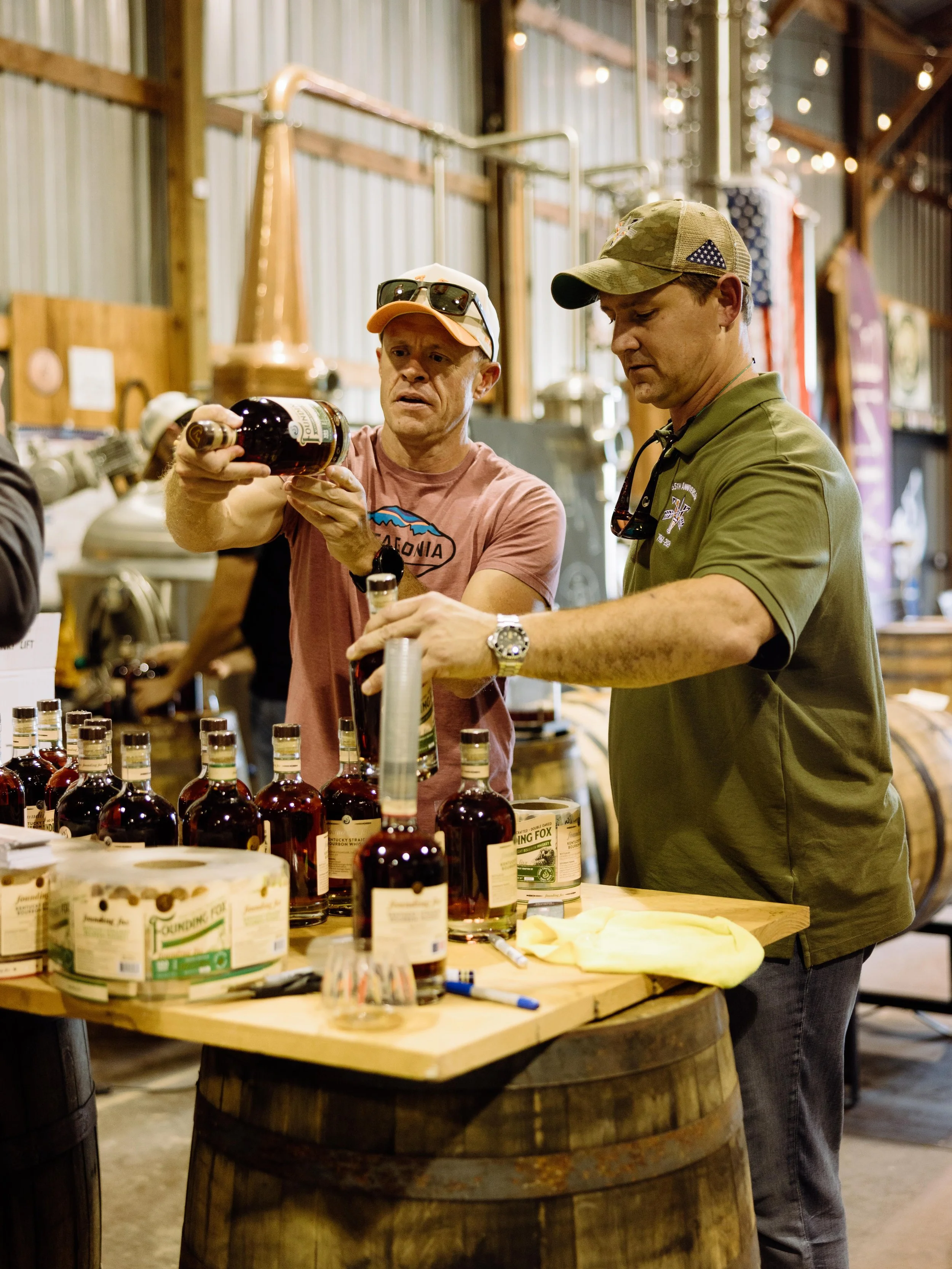 Two men at a table with bottles of bourbon whiskey in a rustic setting with wooden walls and string lights.