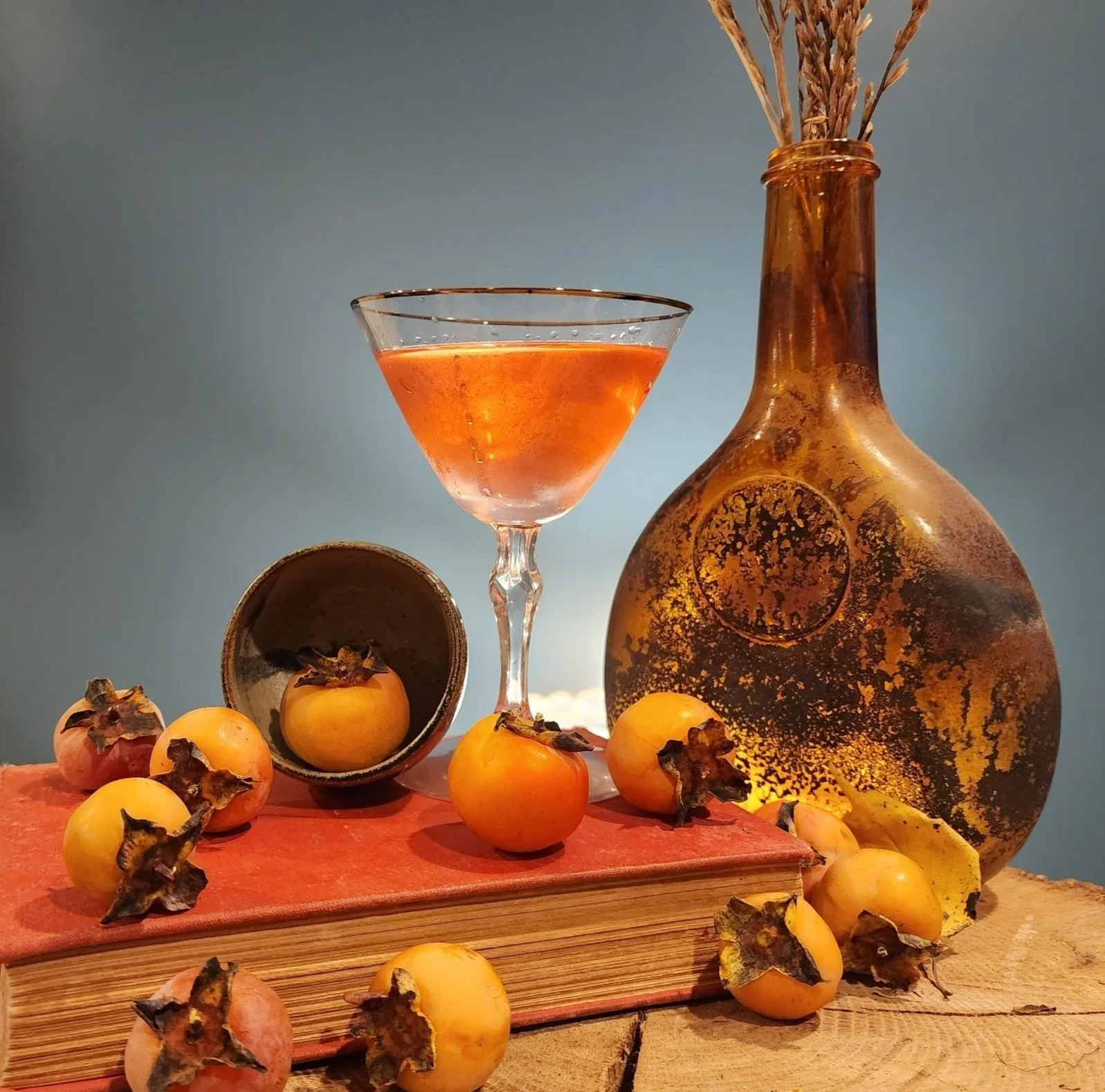 A decorative still life composition featuring a glass of orange-colored drink, a large amber glass vase with dried plants, a wide-open metal container with dried persimmons, and fresh persimmons scattered on a wooden surface with a soft blue-gray background.