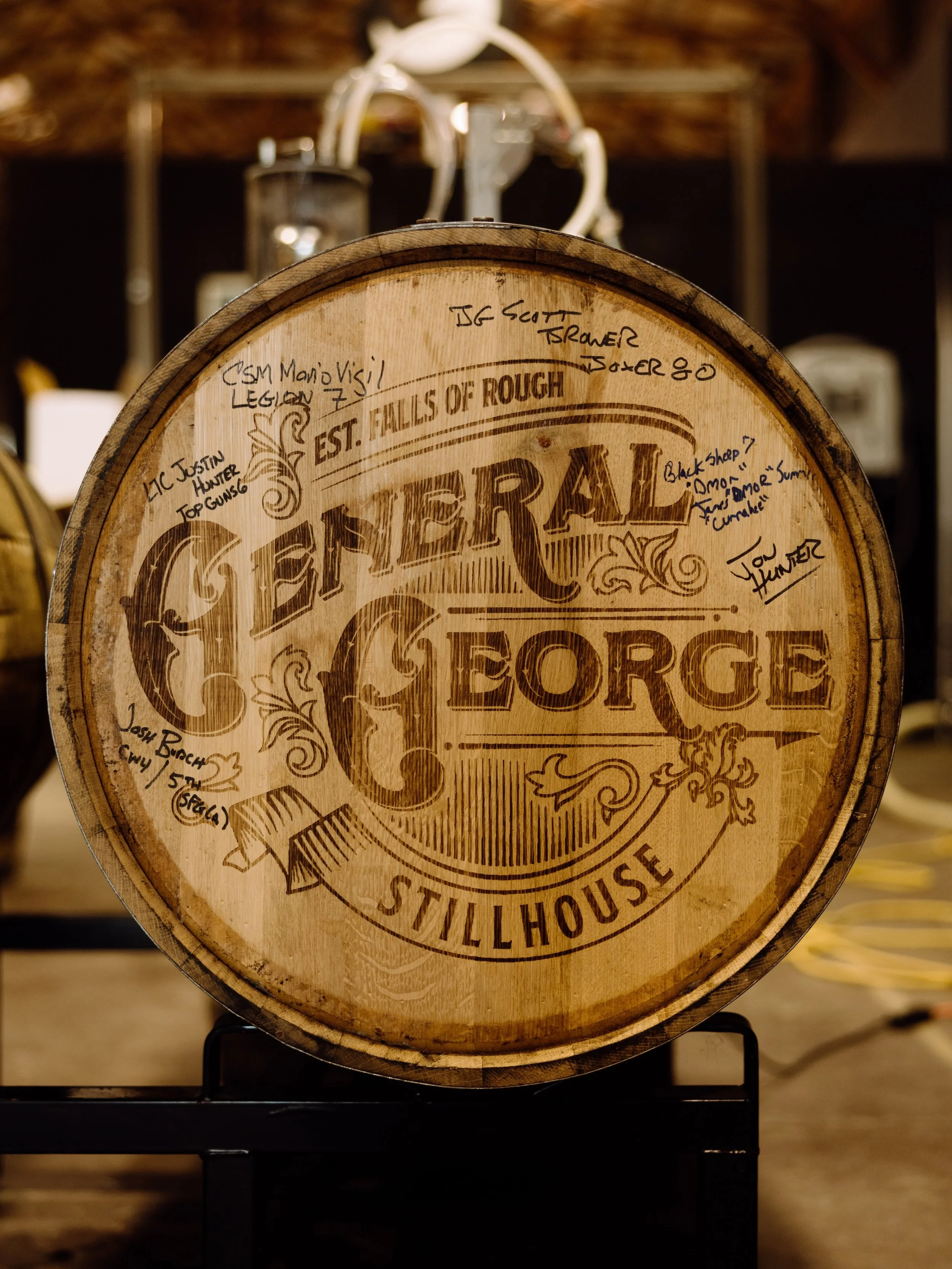 A wooden barrel with engraved text and signatures on the top, displayed on a black stand in a store or brewery setting.