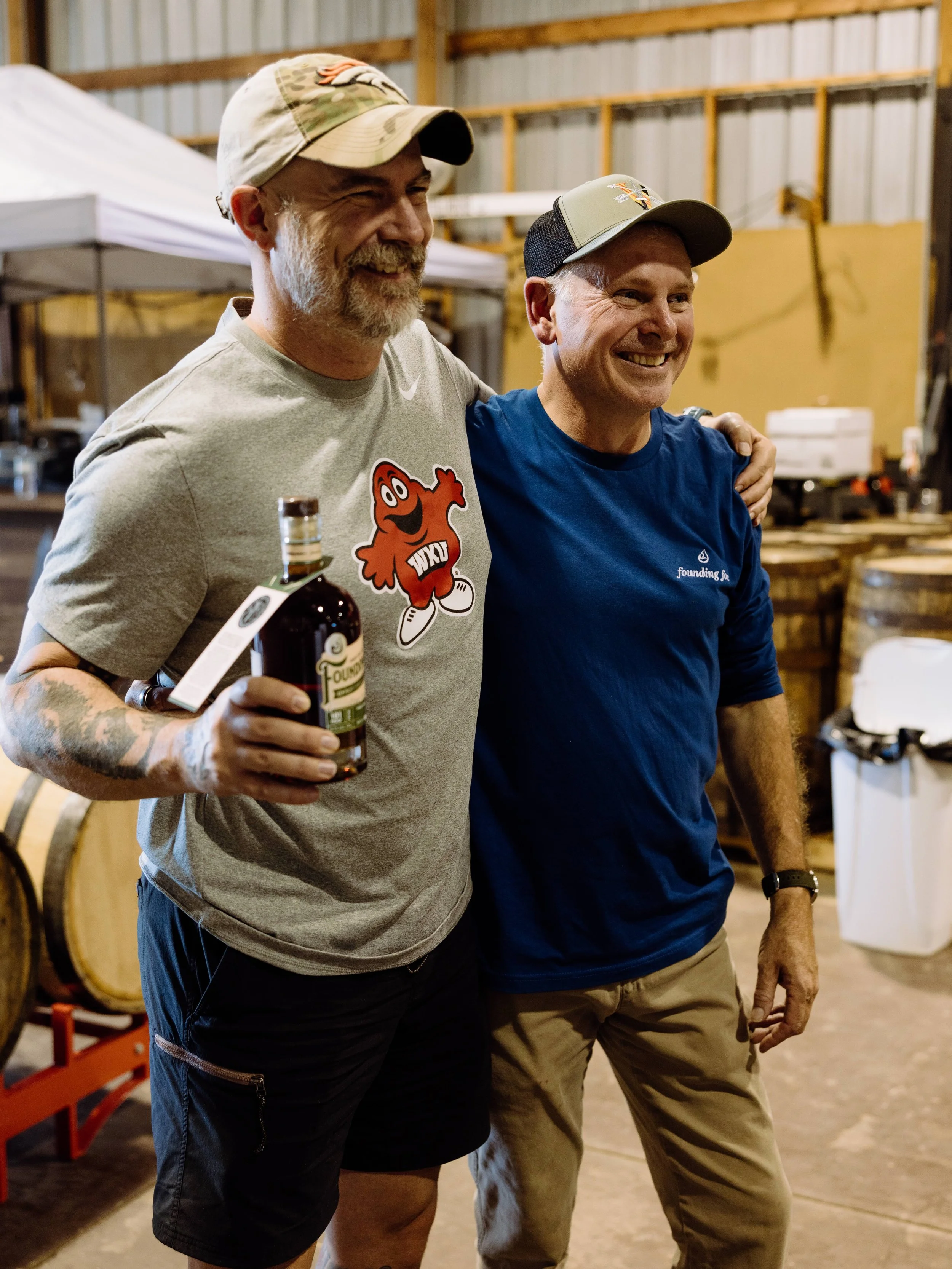 Two smiling men with arms around each other, standing in a rustic barn or warehouse with wooden and metal walls, holding a bottle of alcohol.