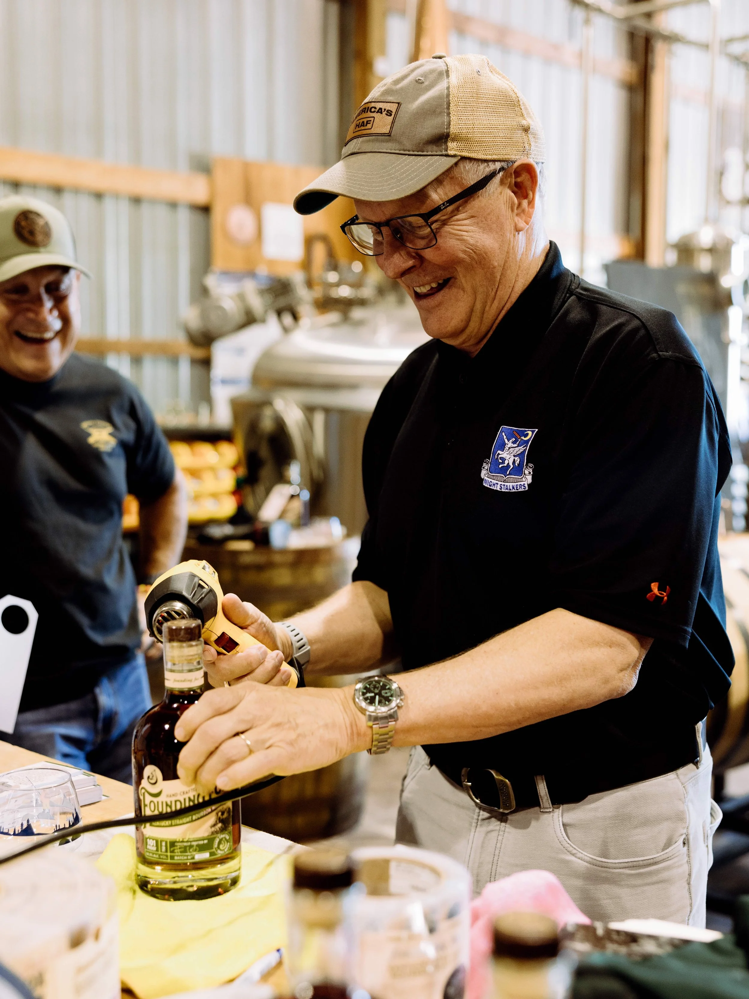 Two smiling men at a bar or kitchen counter, with one man handing a bottle of liquor to the other. The man receiving the bottle is wearing glasses, a beige cap, and a black polo shirt with a patch. The background shows a rustic wooden interior with metal panels.