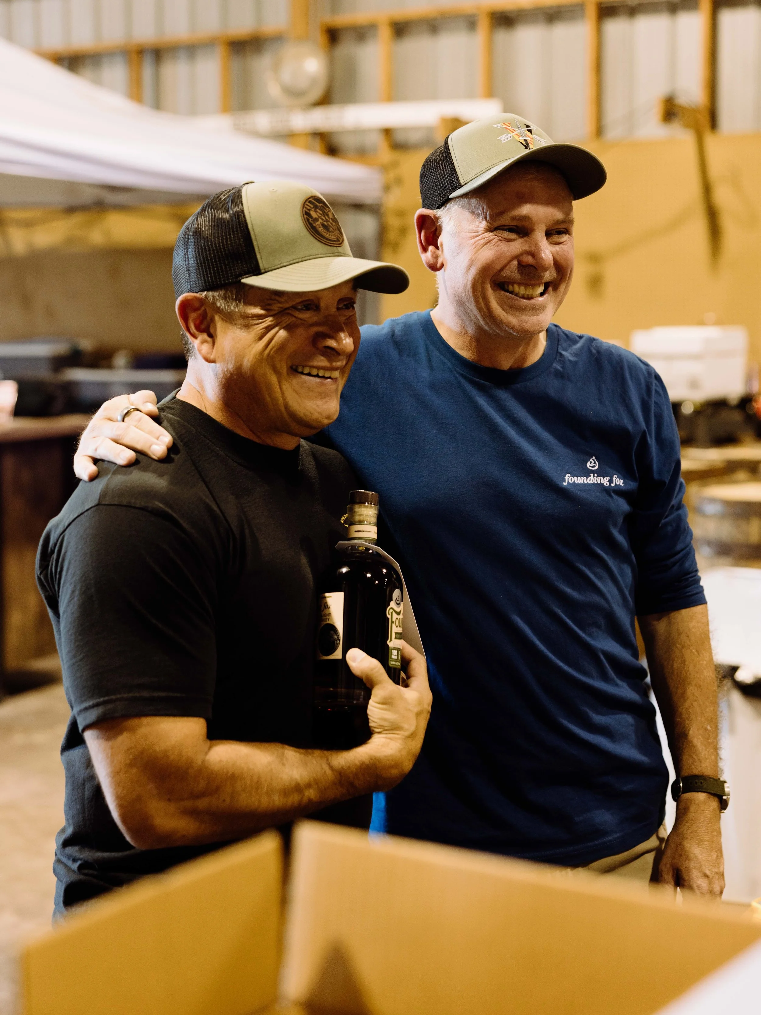 Two smiling men in casual clothing and trucker hats are standing close together, one holding a bottle of Founding F...