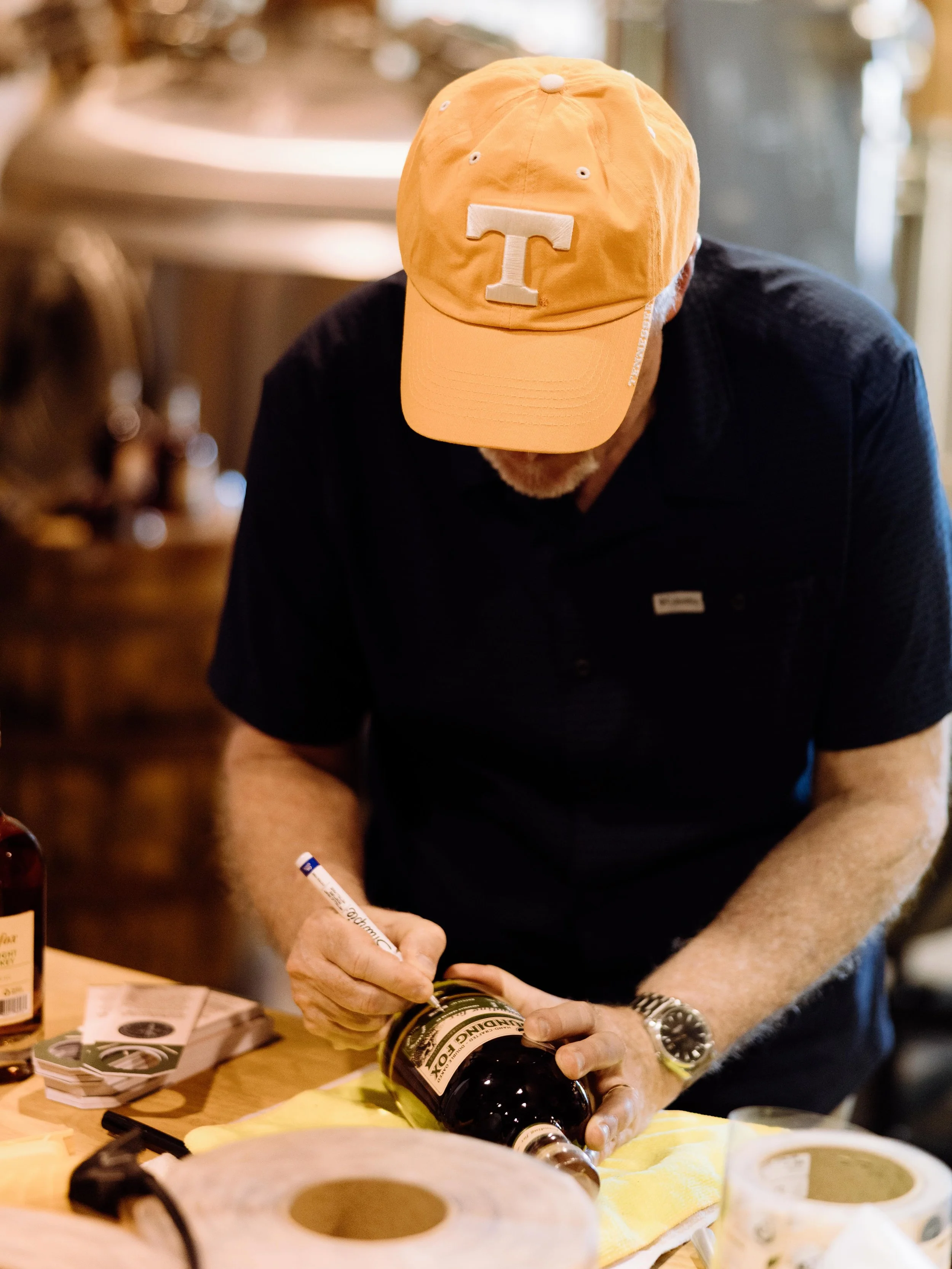 Close-up of a man wearing an orange baseball cap with a white letter 'T' on the front, signing a bottle of Beam Porter at a table with various items and papers.