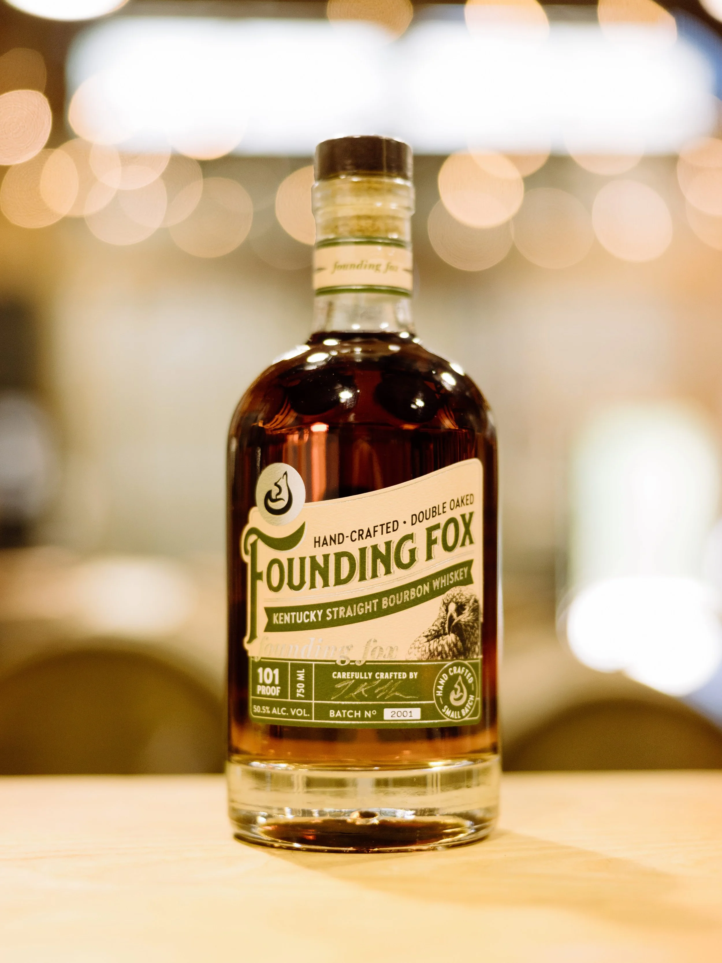 A close-up of a bottle of Founding Fox Kentucky Straight Bourbon Whiskey, with a blurred background of circular lights. The label shows details about the whiskey, including proof, alcohol content, batch number, and branding.