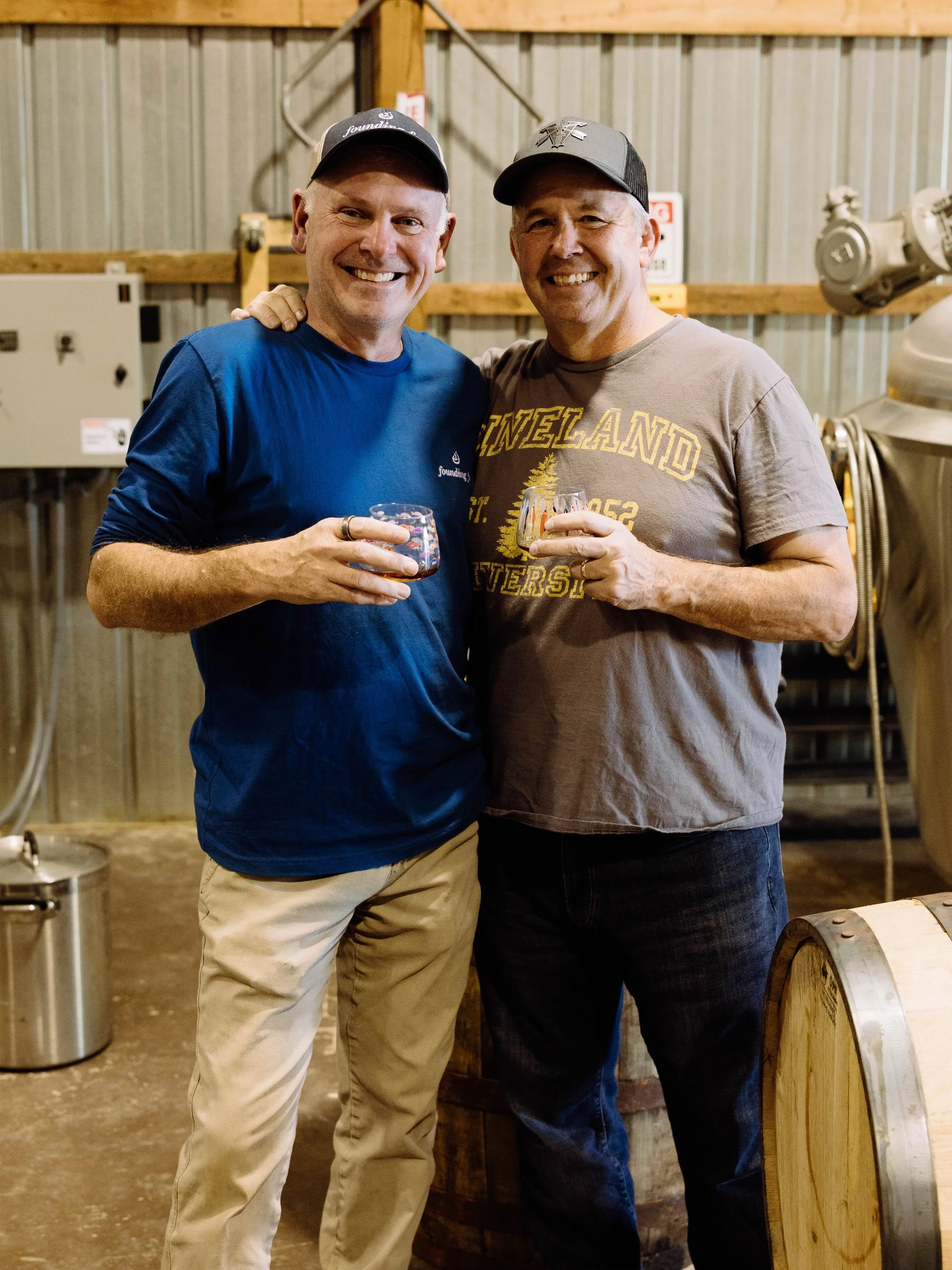 Two smiling men holding glasses of drinks, standing close together inside a rustic wooden and metal building, possibly a brewery or winery, with equipment and barrels in the background.