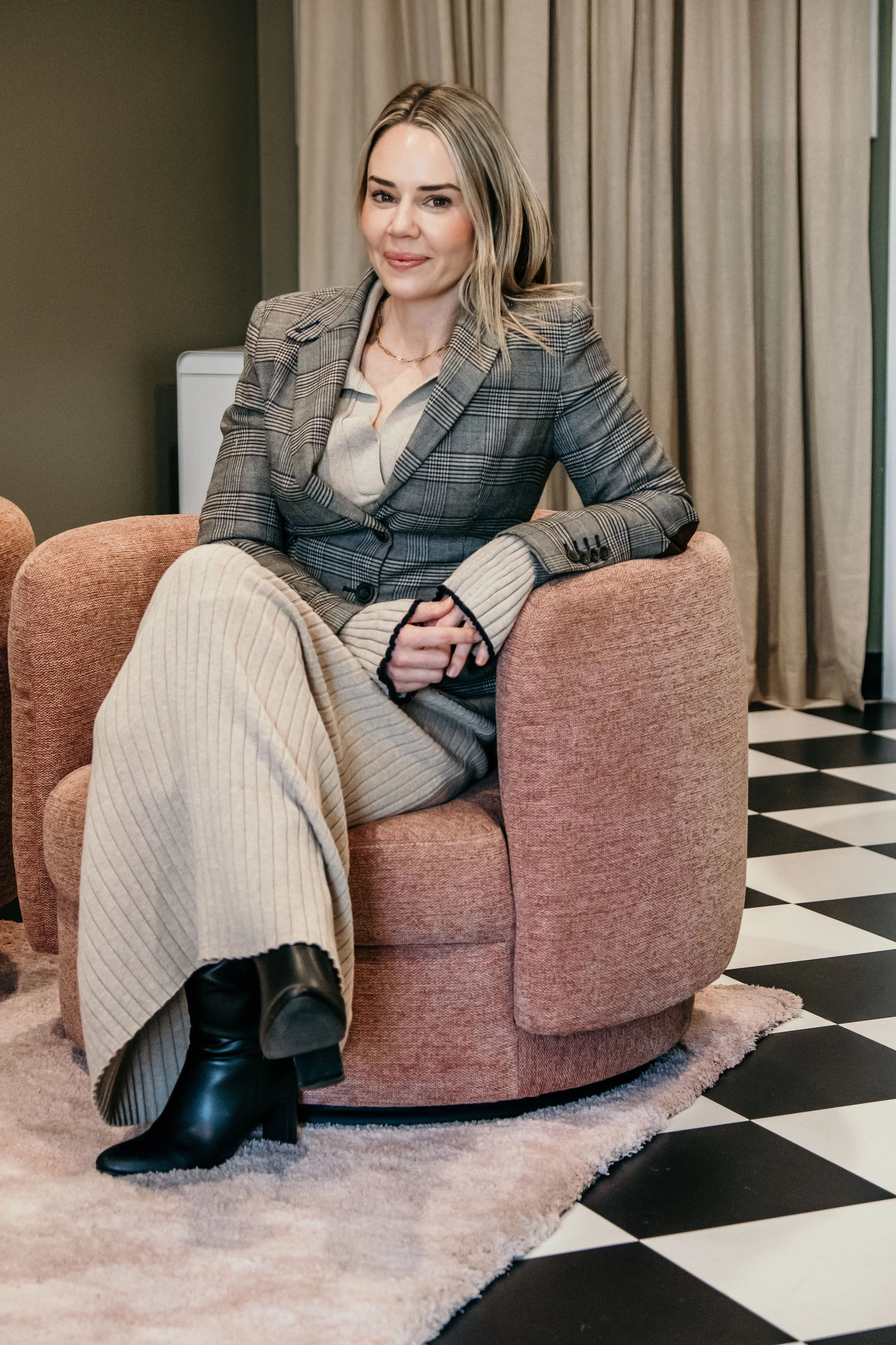 A woman with blonde hair sitting on a pink armchair in a room with black and white checkered flooring and beige curtains. She is wearing a plaid blazer, beige ribbed pants, and black boots, smiling at the camera.