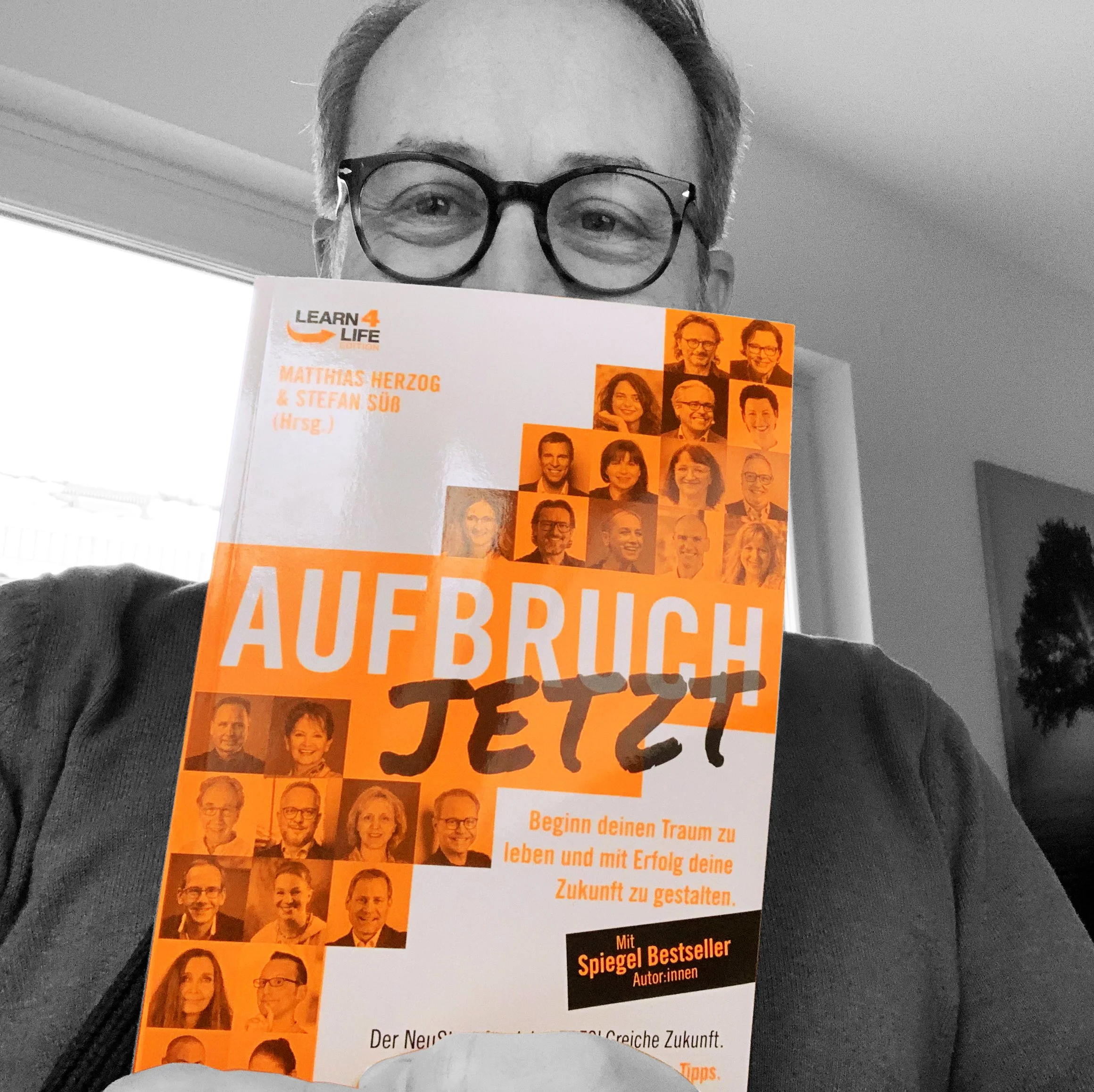 Person mit Brille hält ein Buch mit dem Titel 'AUFBRUCH JETZT' in orange, dessen Cover Porträts mehrerer Menschen zeigt. Autorennamen und das Logo 'LEARN4LIFE' sind ebenfalls sichtbar.