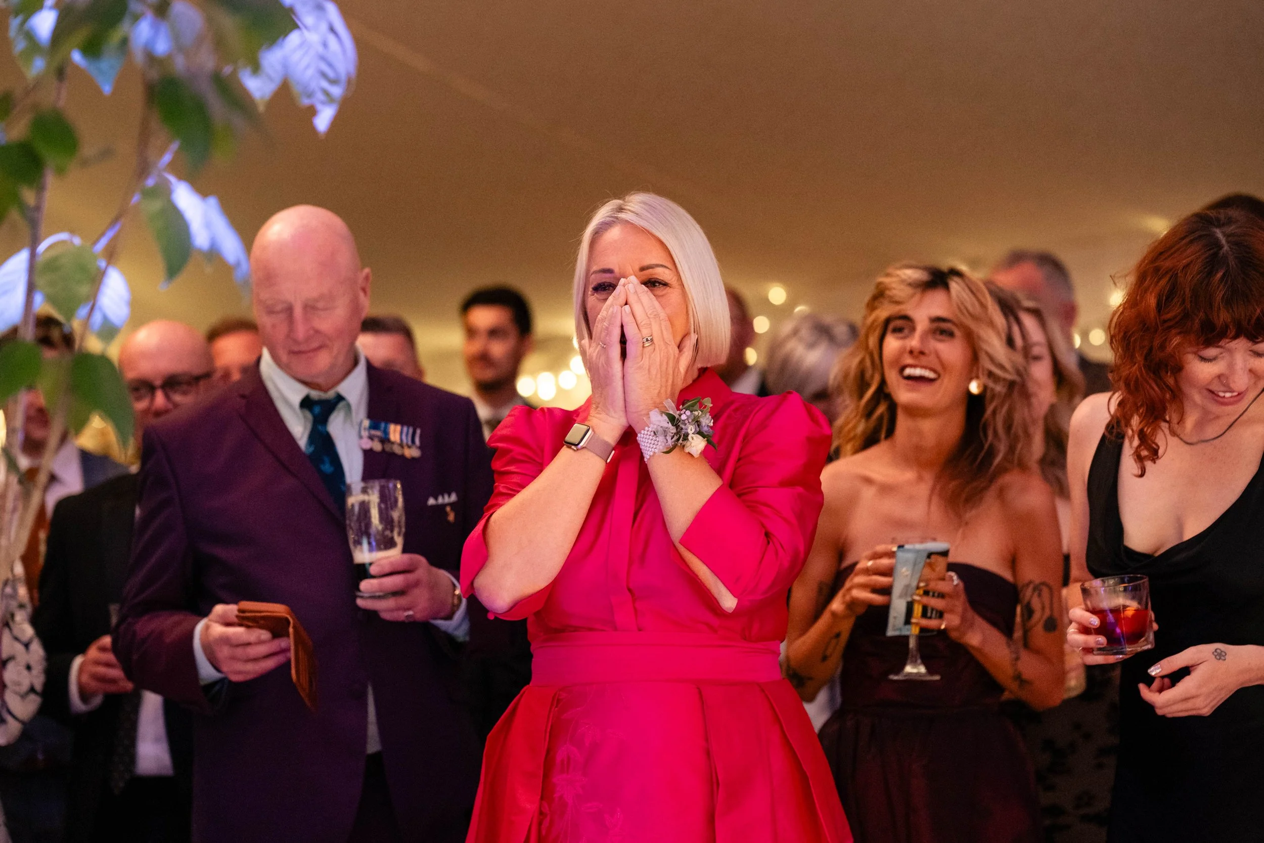 A woman in a bright pink dress is covering her face with her hands, appearing emotional at a social gathering. She is surrounded by people smiling and watching her, some holding drinks. A man in a suit with medals and glasses, and women in elegant dr
