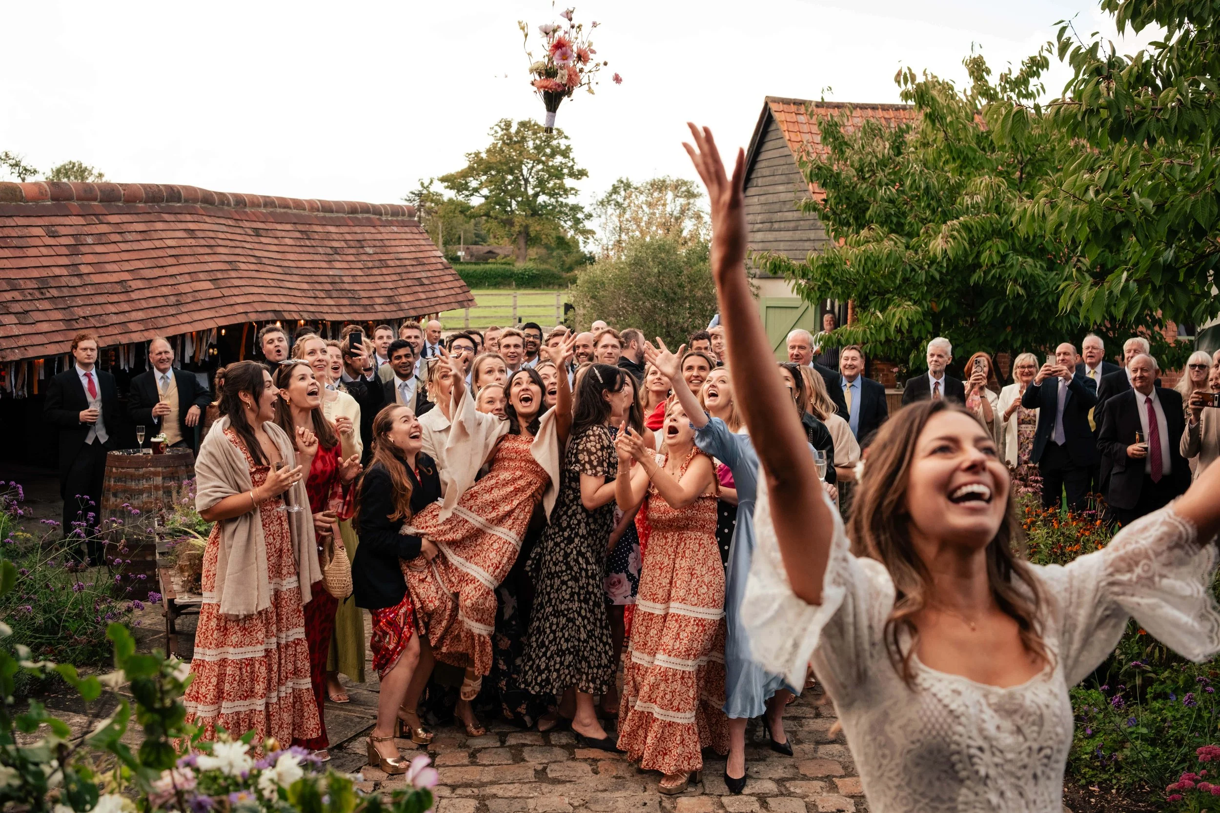 A joyful outdoor celebration with a large group of people, many women in long dresses, cheering and smiling with one woman in the foreground raising her arms triumphantly during daytime in a rustic garden setting.