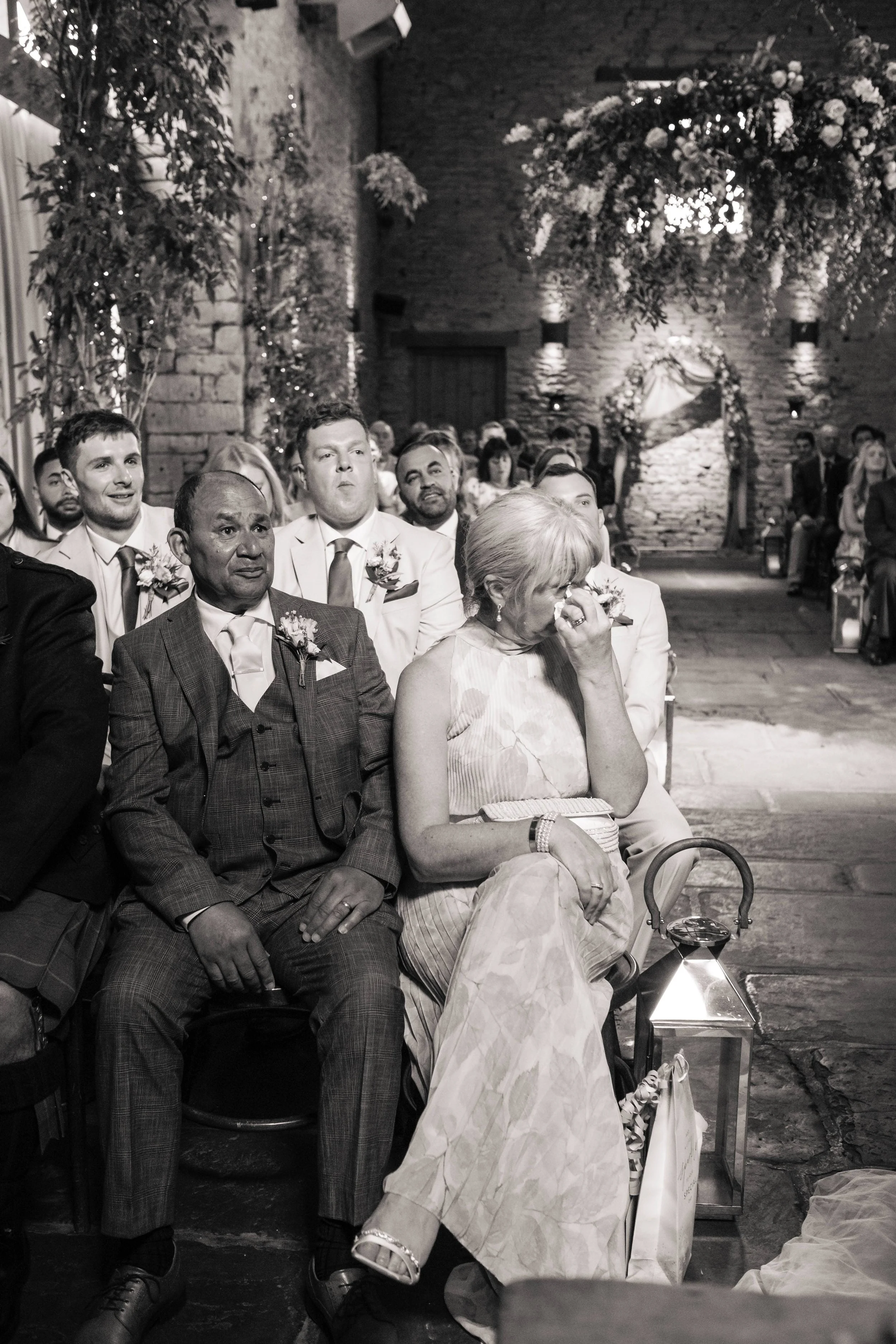 A group of people attending a wedding ceremony, with some visibly emotional. The setting includes a stone wall and floral arrangements.
