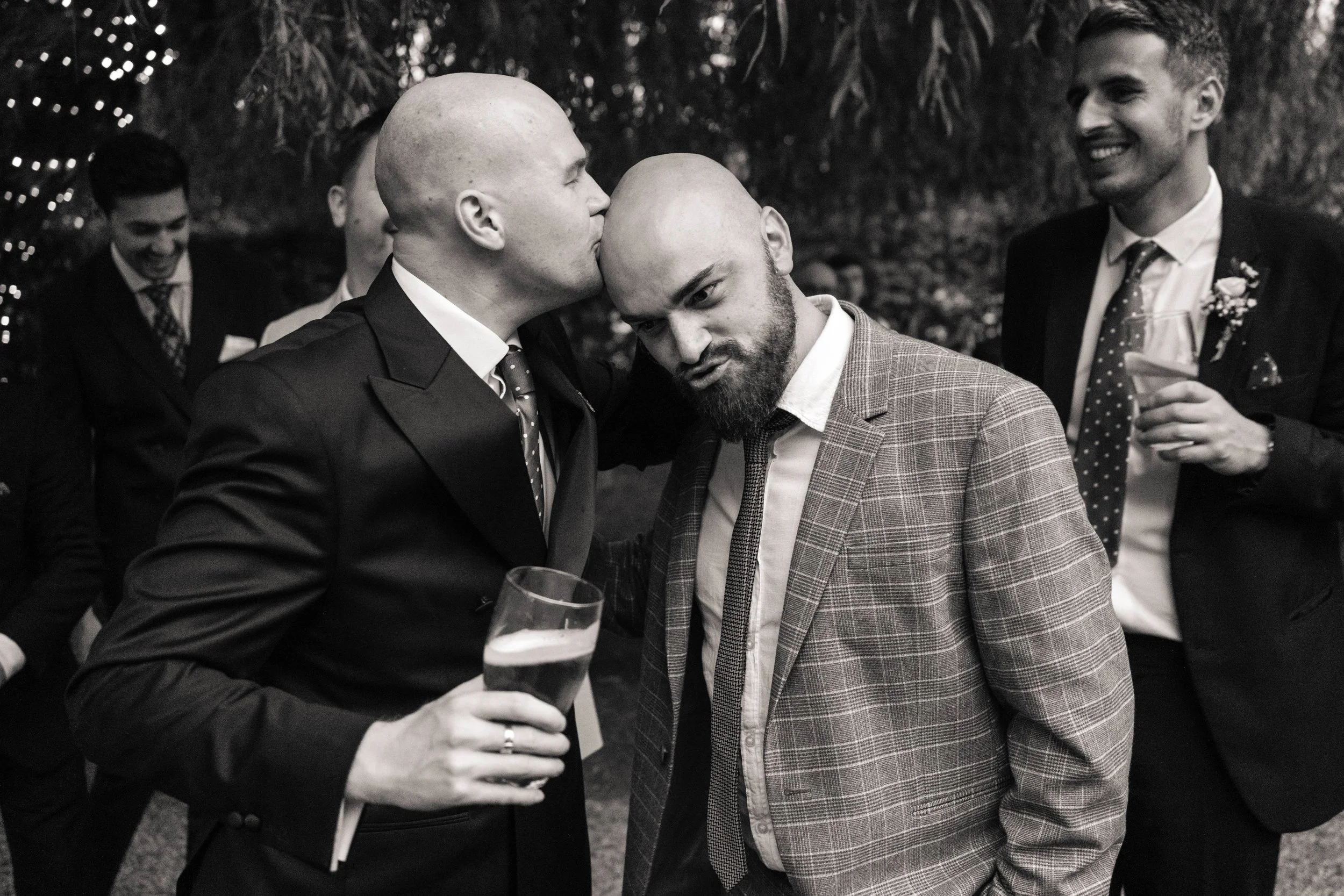 The groom with a shaved head and suit kisses a wedding guest with a beard and plaid blazer, who looks surprised, while two other men in suits laugh in the background, one holding a glass of wine.