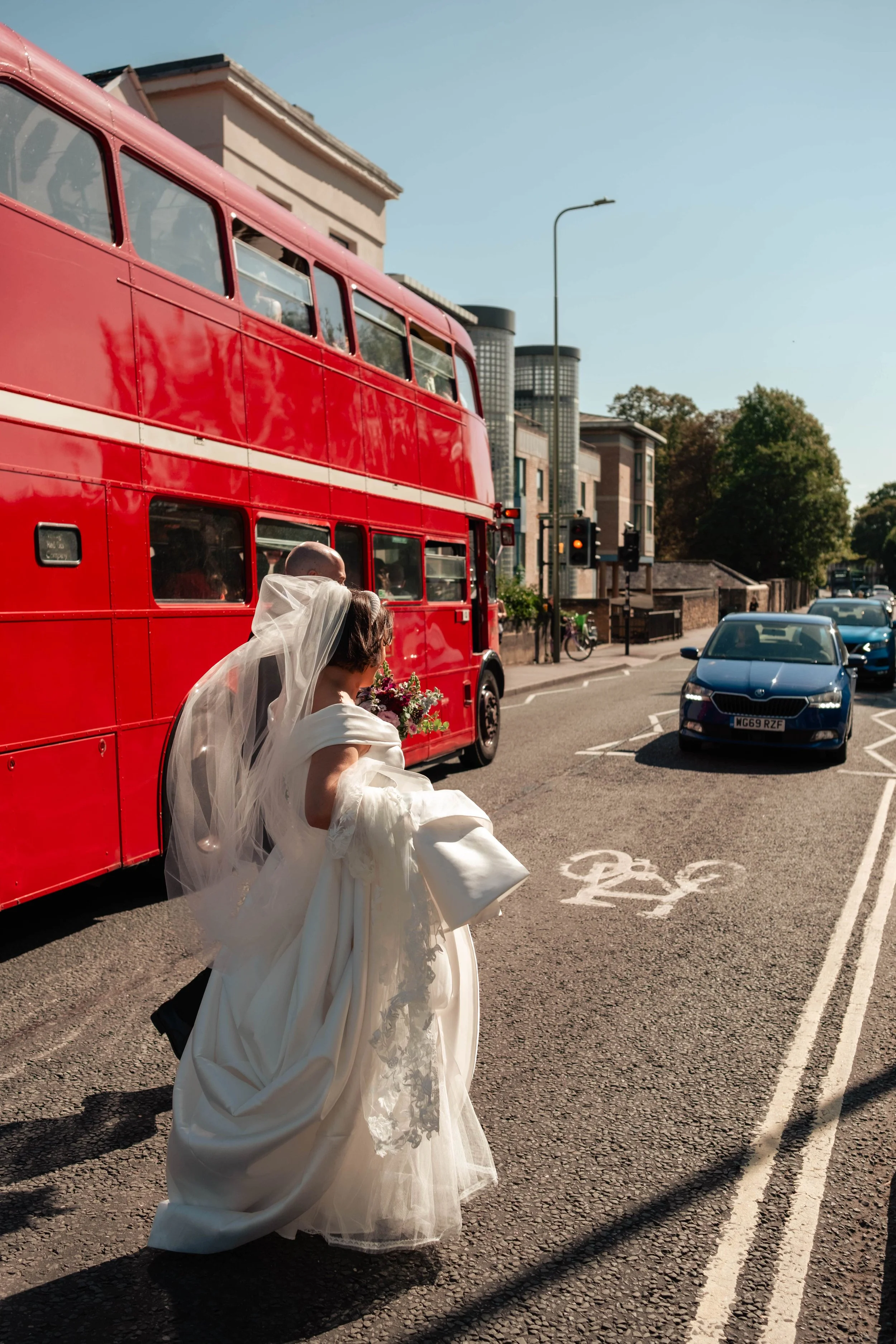 A bride in a white wedding dress and veil is holding a bouquet and walking across a city street near a red double-decker bus and cars, with buildings and a bicycle in the background.