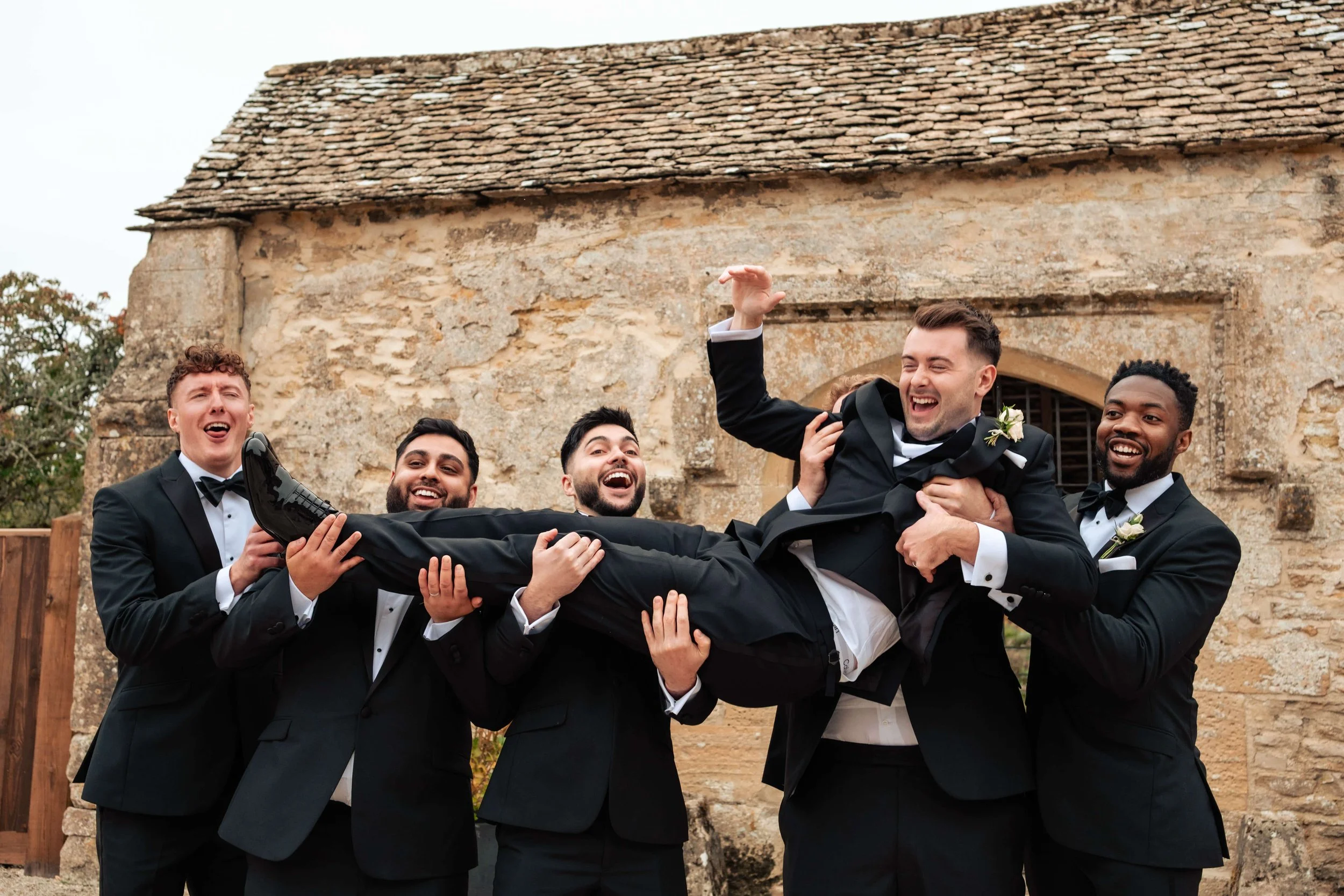 Groomsmen in tuxedos lifting the groom in a tuxedo horizontally while smiling and celebrating outdoors.