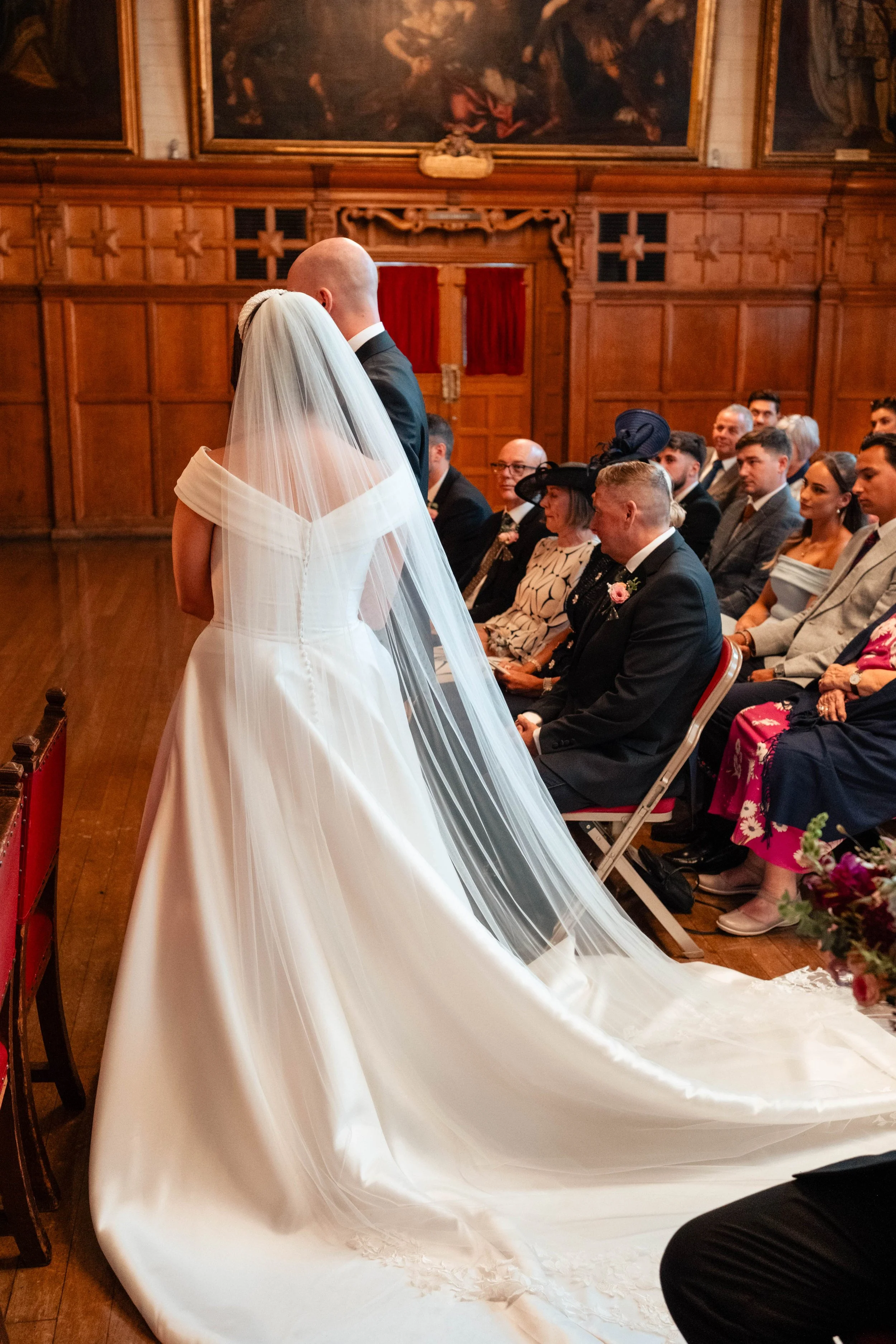 A bride and groom standing closely during their wedding ceremony in a wood-paneled room with guests seated, a large painting above, and red curtains in the background.