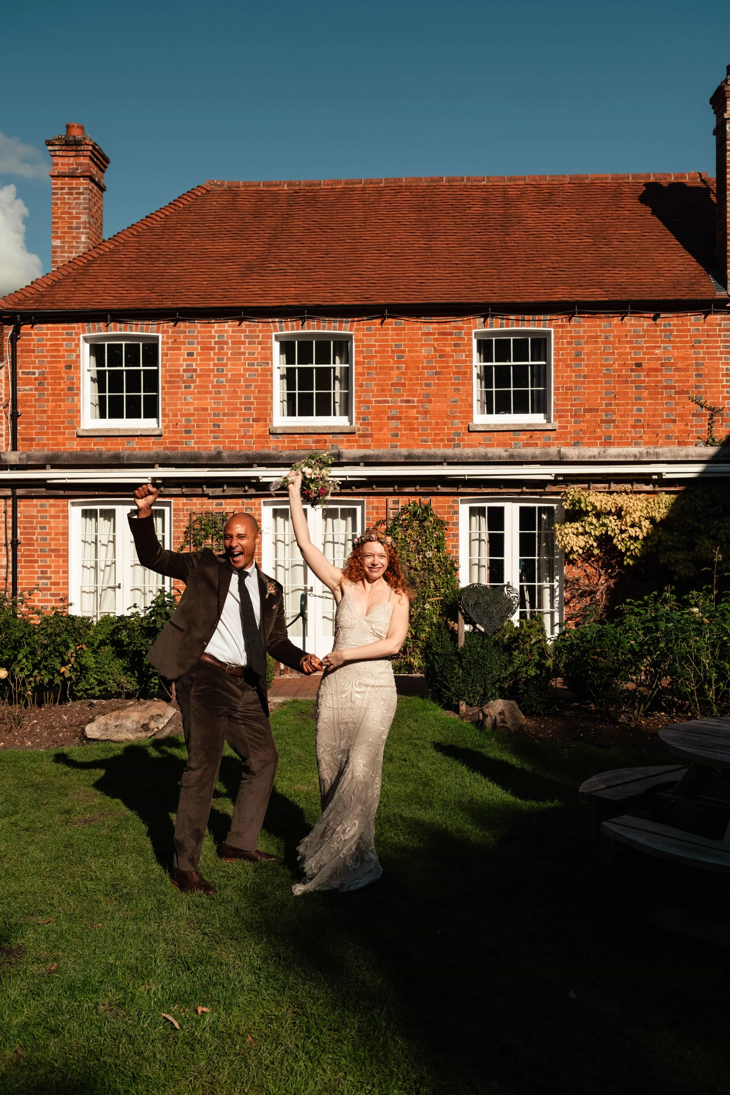 A happy couple celebrating, with the man in a suit and the woman in a wedding dress, holding a bouquet of flowers outdoors in front of a brick house.