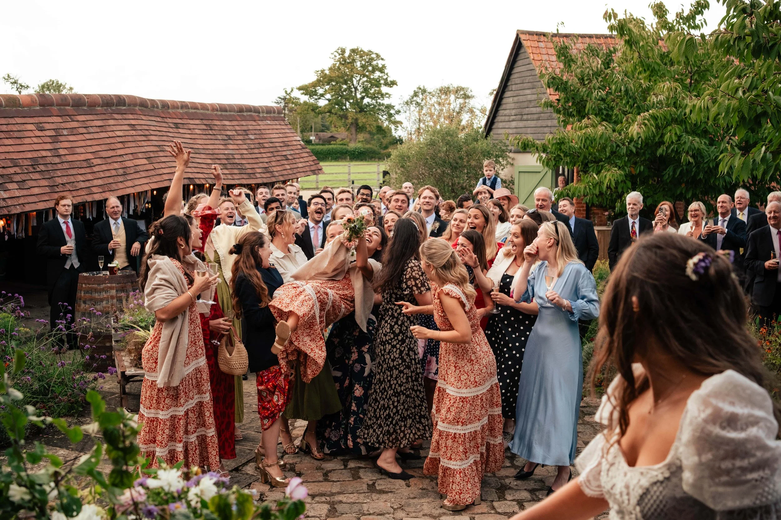 A large group of people celebrating outdoors on a rustic brick patio, with some holding drinks and one woman being lifted in the air, surrounded by trees and farm buildings.