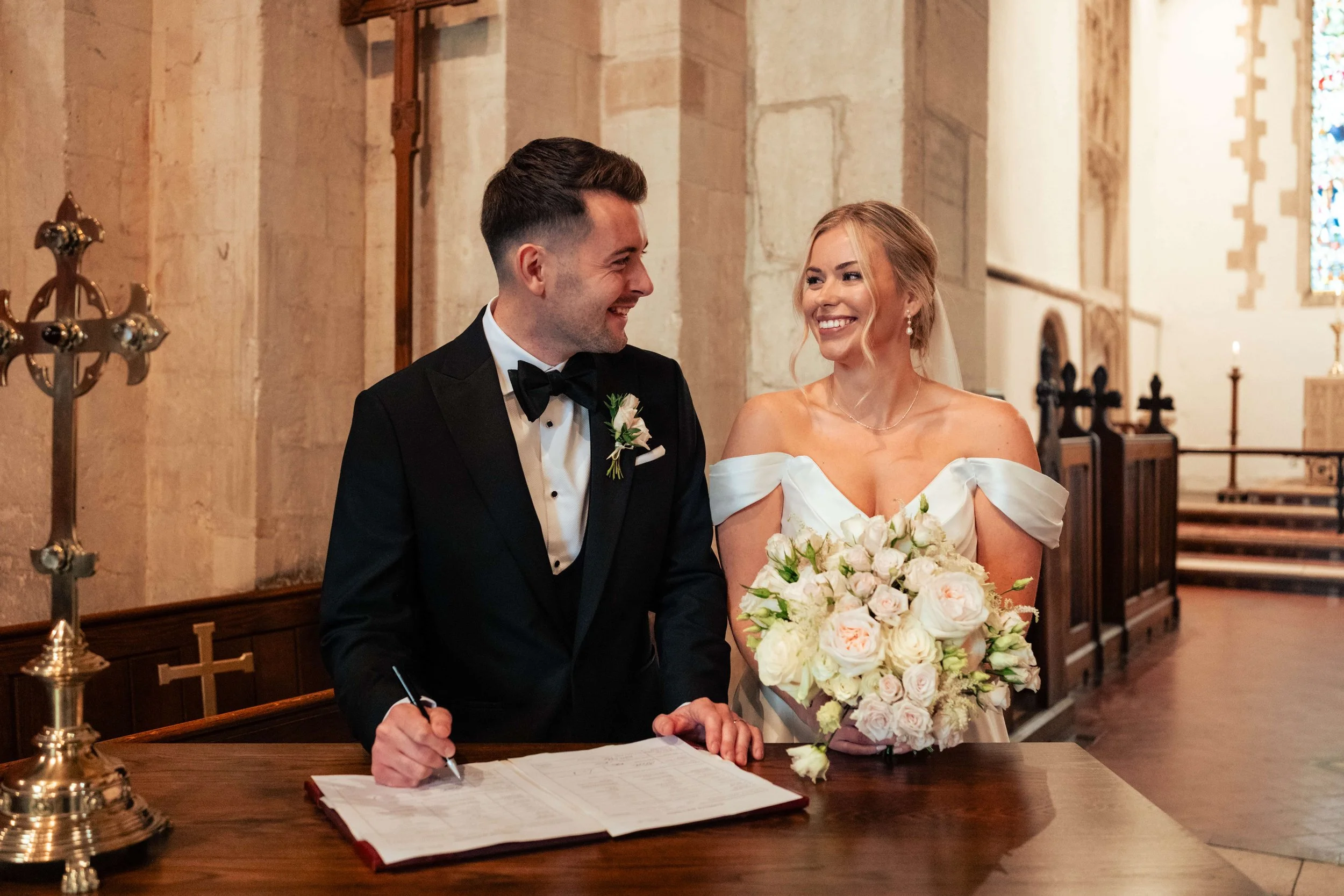 A bride and groom at their wedding ceremony inside a church, smiling at each other. The groom is signing marriage documents while the bride holds a bouquet of white and pink flowers.