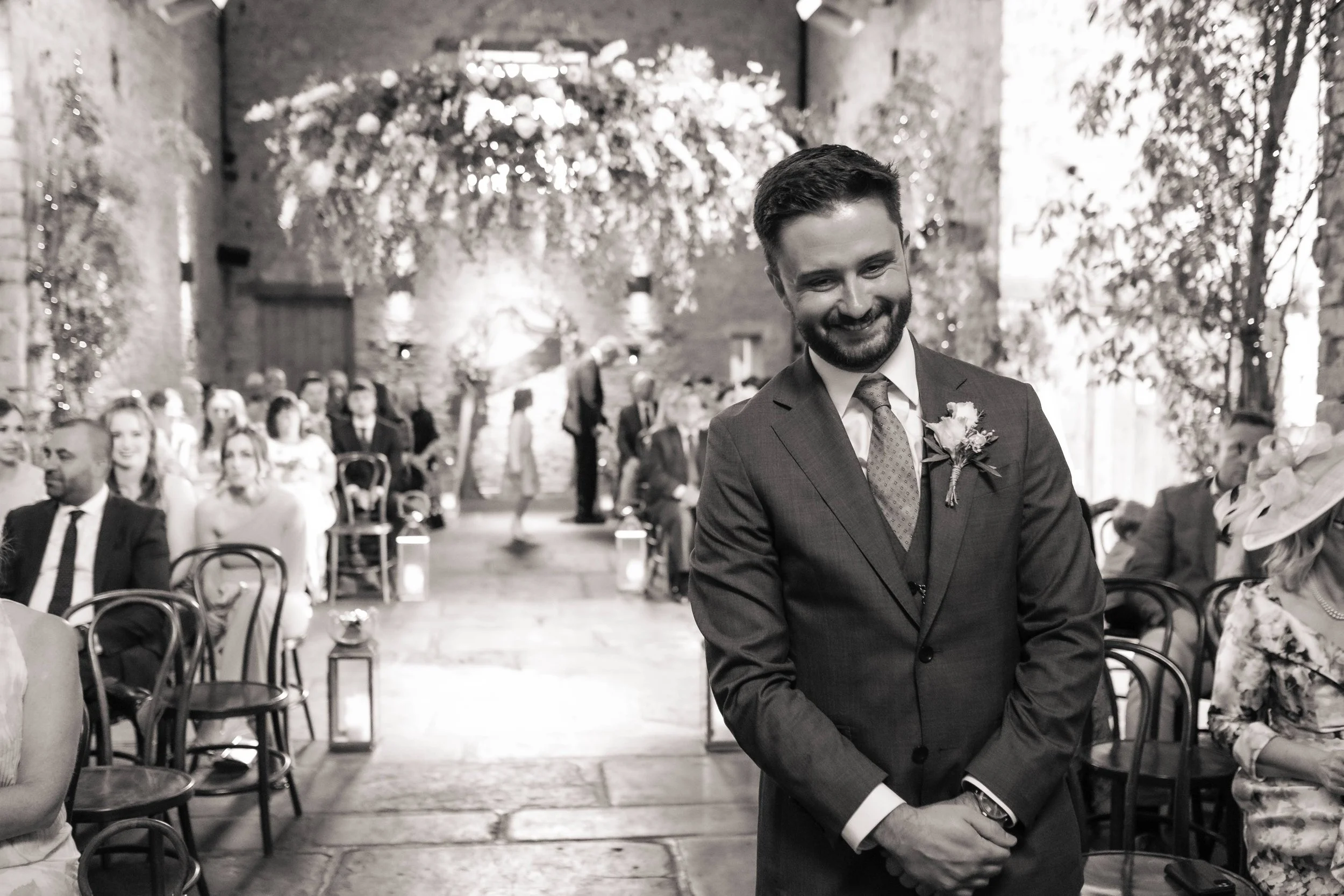 A smiling man in a suit stands with crossed hands during a wedding ceremony. Guests are seated behind him, and the venue is decorated with hanging flowers, trees, and candles.