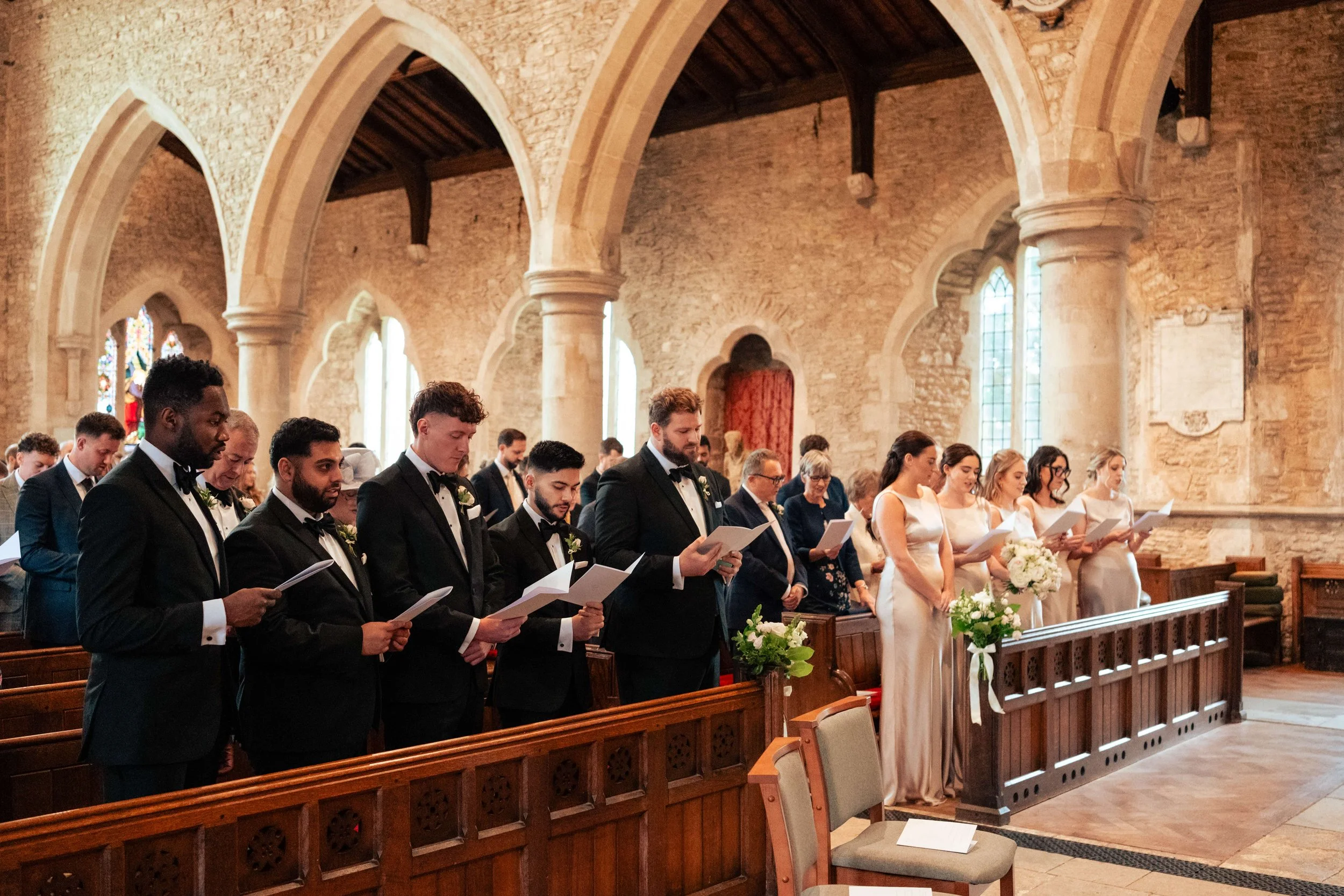 A wedding ceremony taking place inside a historic stone church with stained glass windows. The bride and groom, along with the bridal party and guests, are standing at the altar reading from papers.