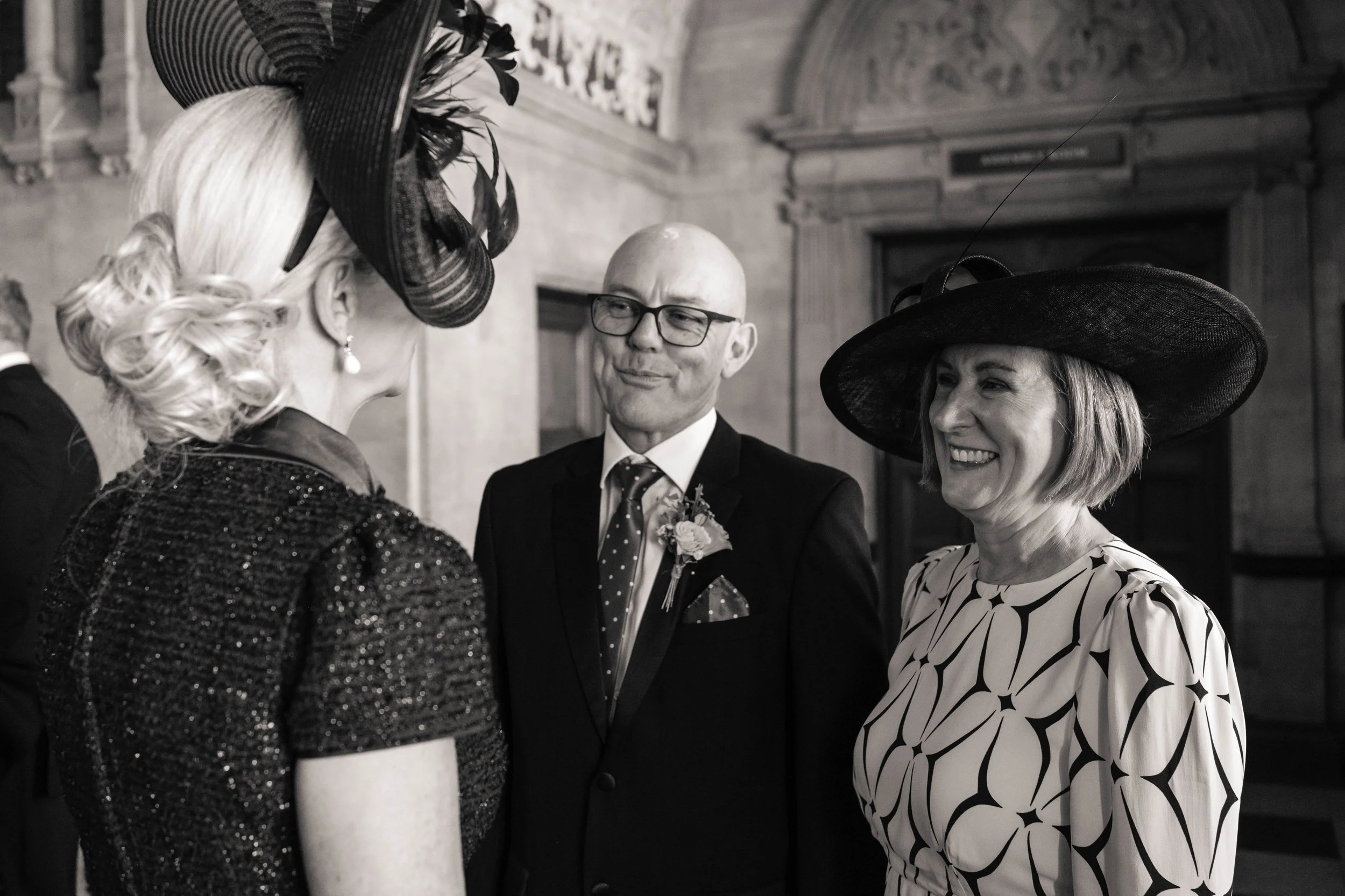 Three people conversing at a formal event, two women wearing large hats, one woman with a patterned dress, and a man in a suit with a boutonnière, in a wood-paneled room.