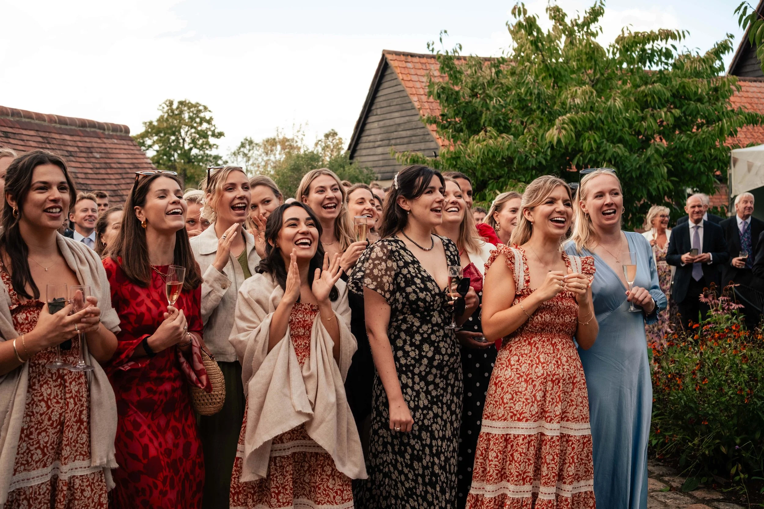 Group of women in colorful dresses at an outdoor celebration, some holding champagne glasses, smiling and laughing.