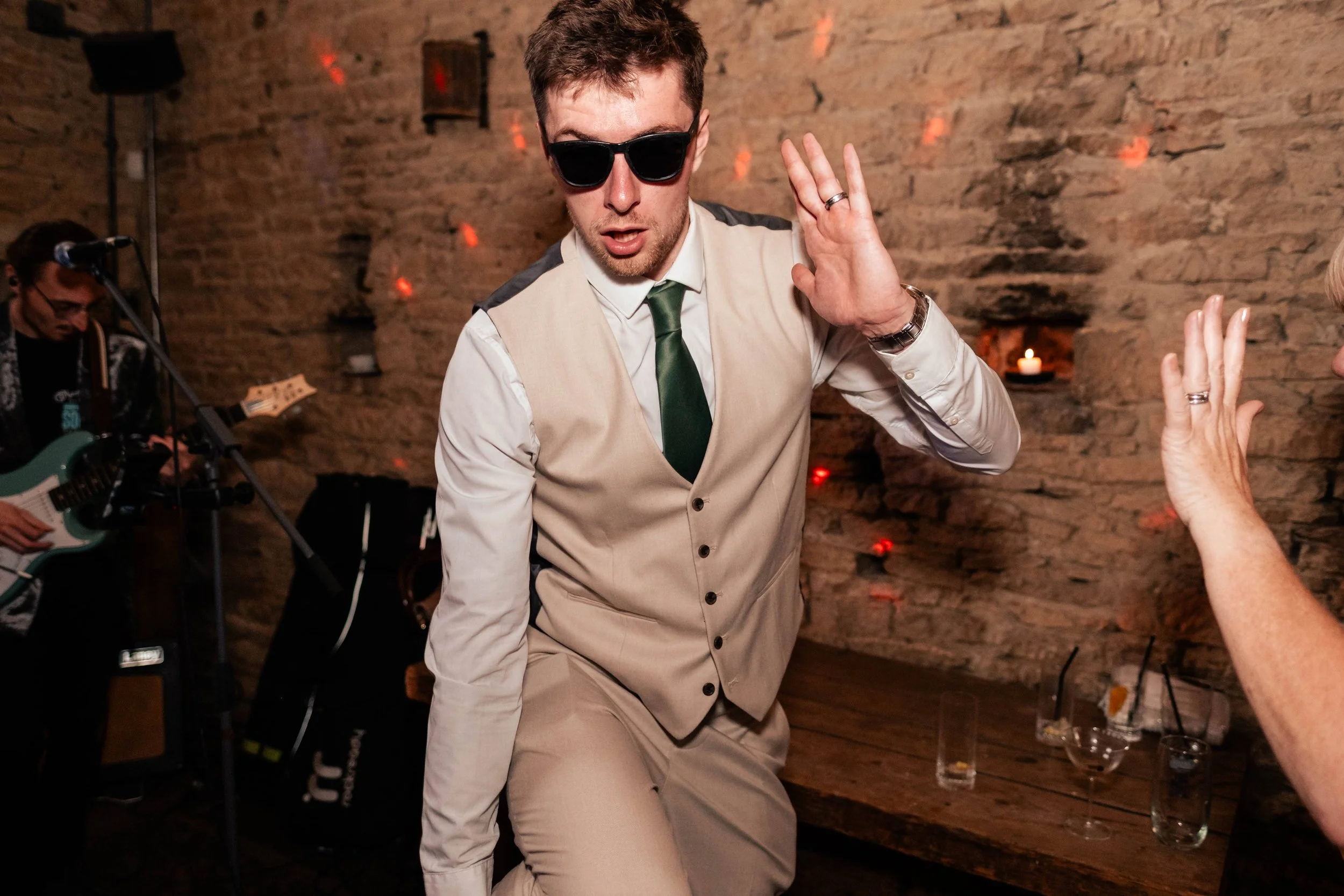 A man in a beige vest and pants, white shirt, green tie, and sunglasses dancing with a woman at a party, with a musician playing guitar in the background and drinks on the table.