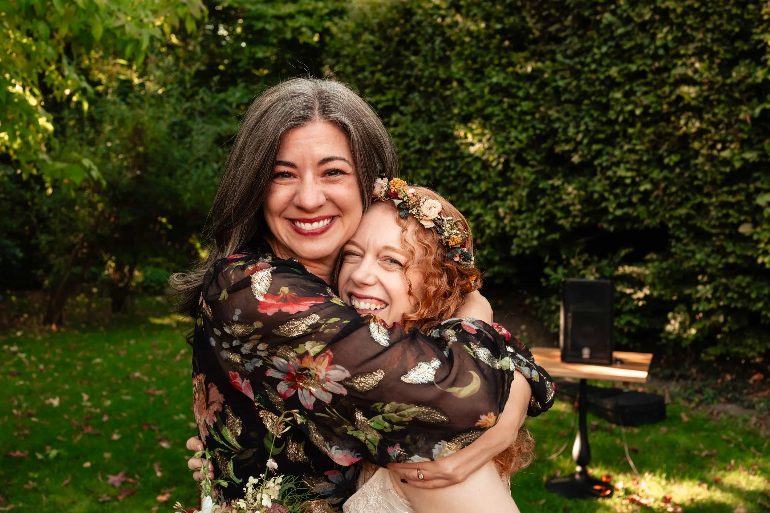 Two women hugging and smiling outdoors, one with dark hair and the other with curly red hair and a floral headpiece, in a garden setting.