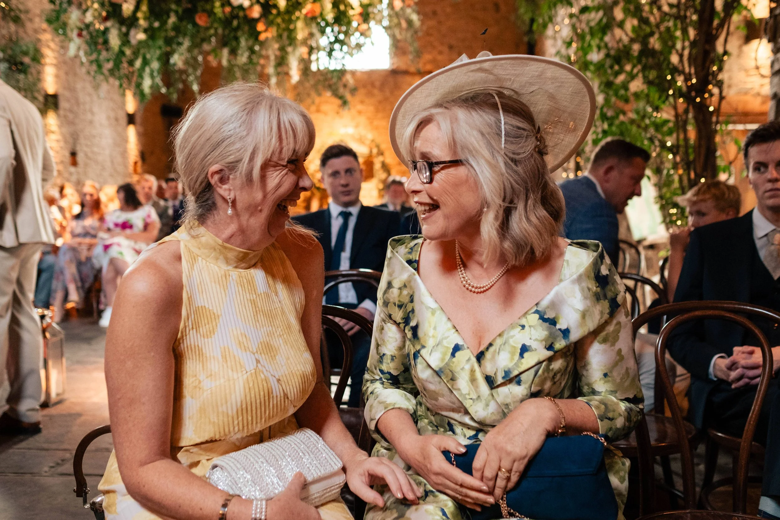 Two women in elegant dresses are smiling and laughing while sitting at a social event. One woman wears a yellow patterned dress and is holding a beaded clutch; the other wears a floral dress with a pearl necklace and large hat with glasses. People ar