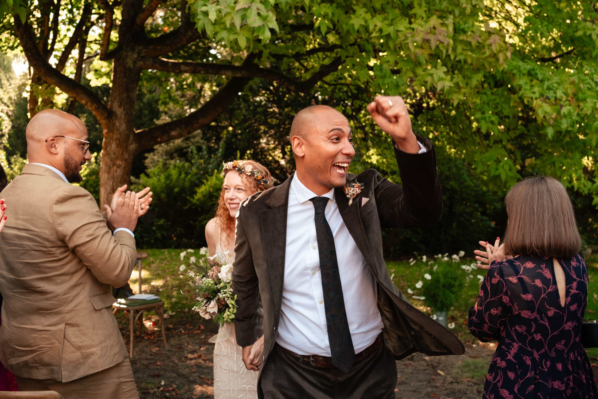A man in a suit celebrating at an outdoor wedding, surrounded by smiling guests clapping and applauding under a large tree.