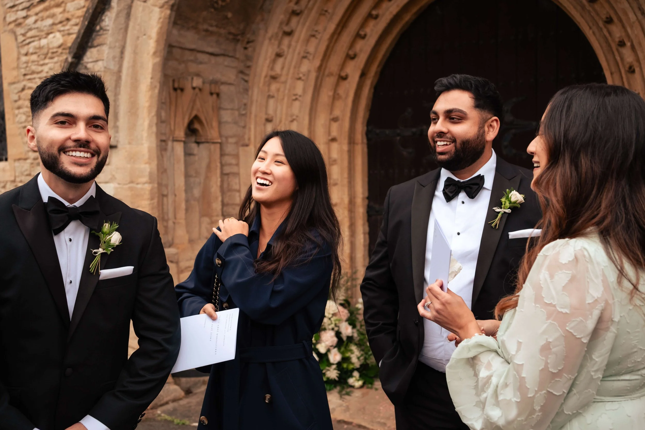 Group of happy people in formal attire at a wedding or celebration outside a stone church with an arched doorway.